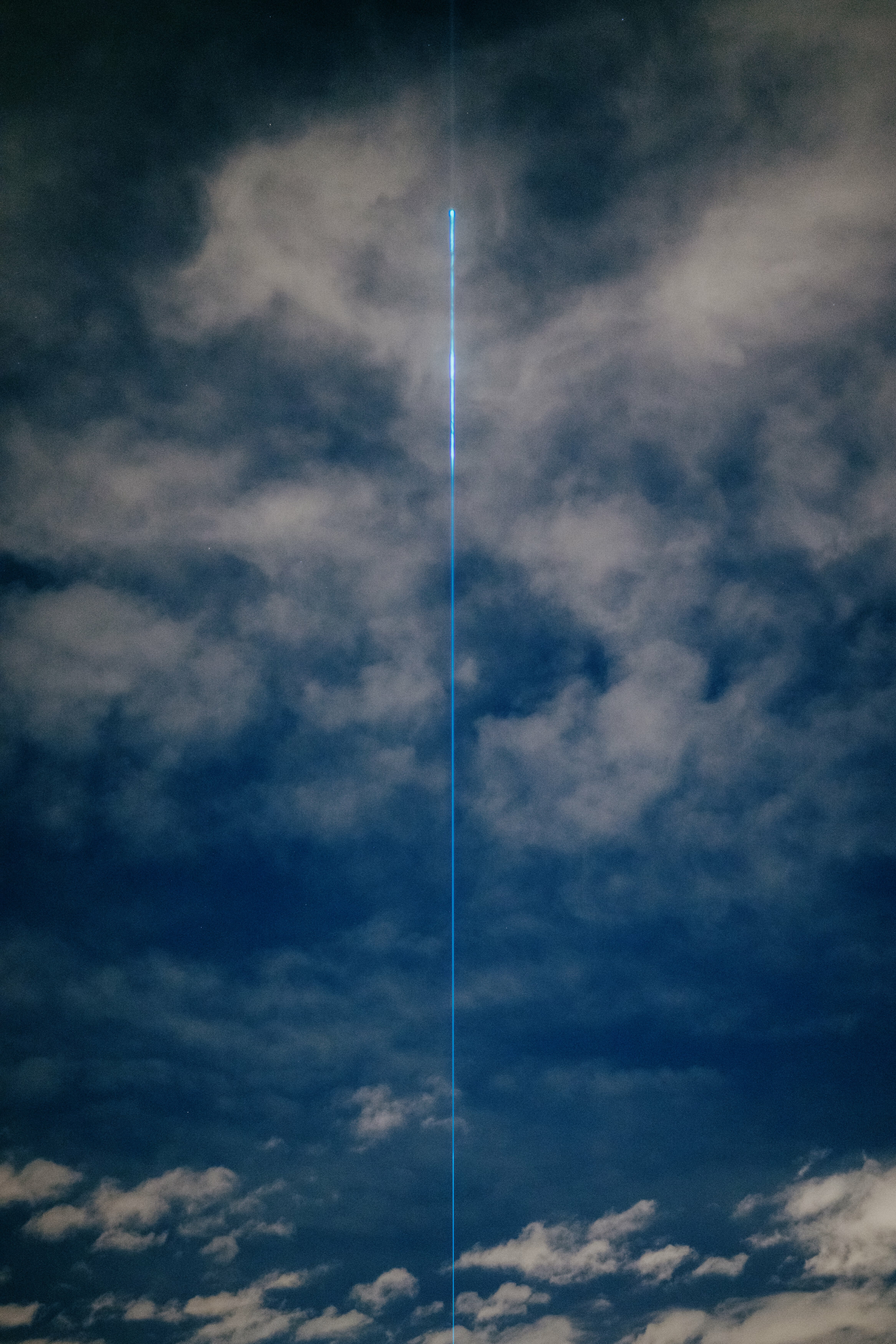 Abstract images clouds at night, a bright streak of light bisects the frame rising up from the middle of the bottom edge and getting brighter as rises before disappearing abruptly behind a cloud.