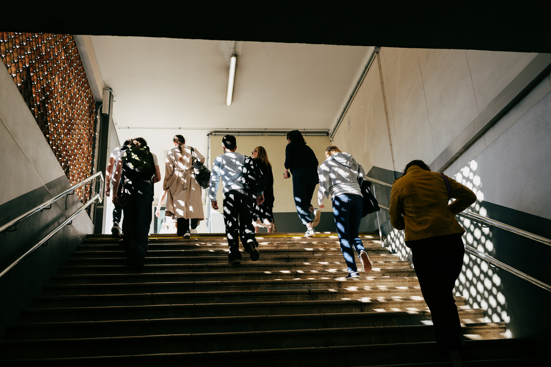 Auto-generated description: A group of people are walking up a staircase in a well-lit indoor space with sunlight casting patterned shadows.