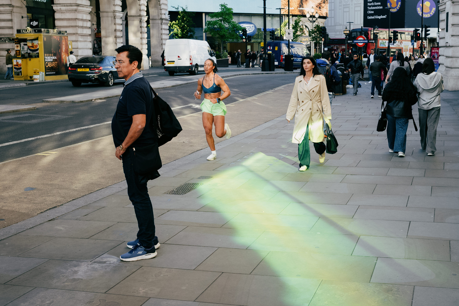 street scene showing some pedestrians walking on a wide pavement. There is an athletic female runner wearing neon green shorts jogging between the pedestrians and what looks like a neon green energy beam emanating from her across the pavement 
