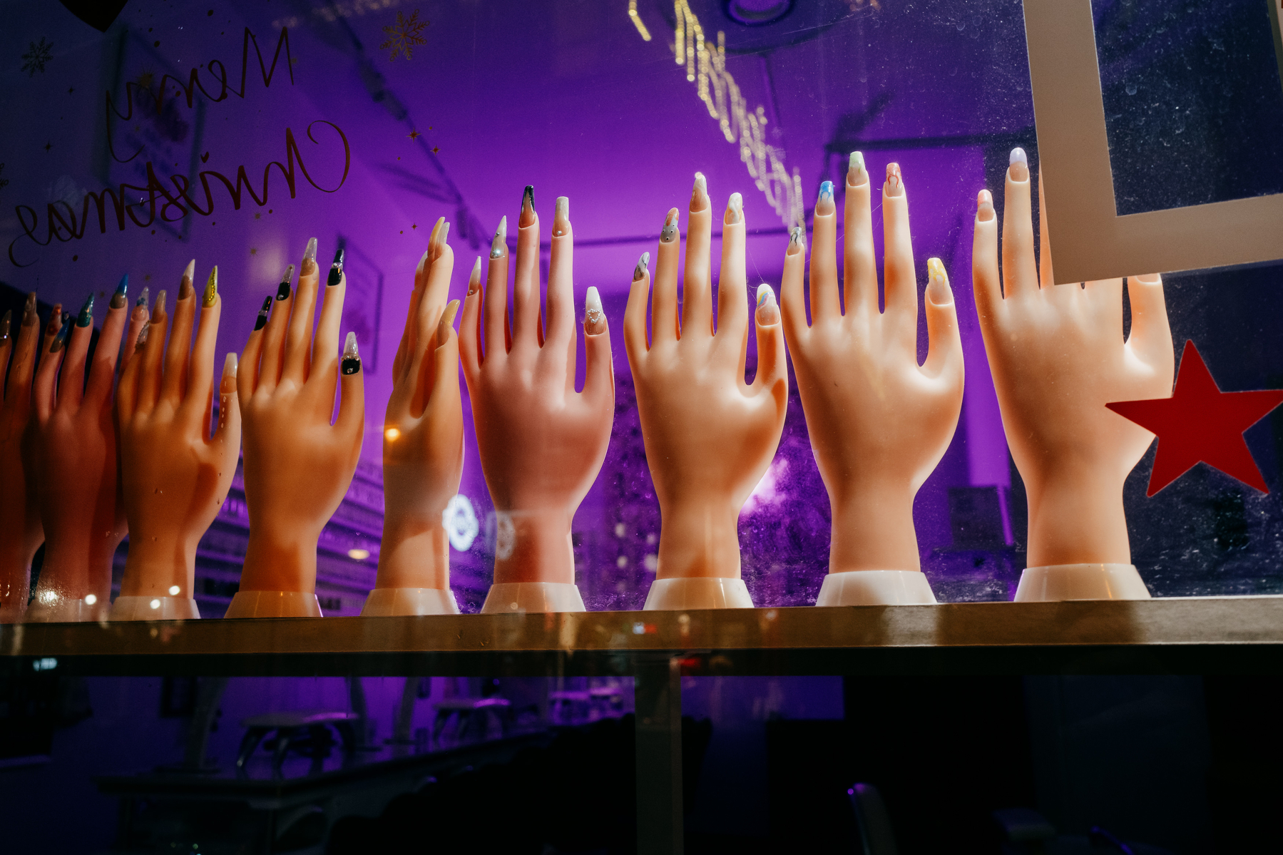 row of mannequin hands in nail salon window with red star stuck on glass at right of frame. The otherwise dark background is lit with the glow of a purple light.