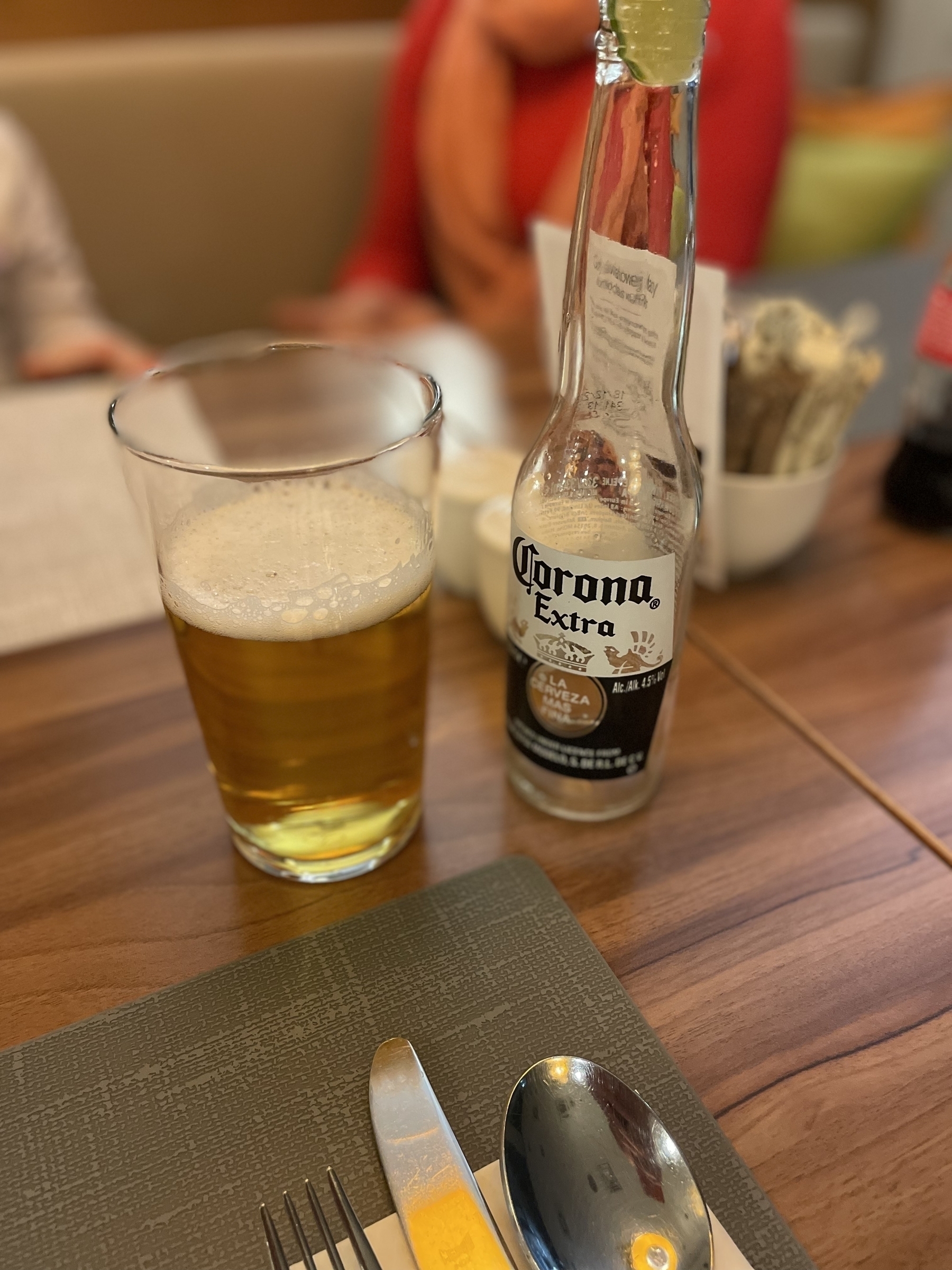 a glass of beer on a table. The beer is Corona. In the background we have a restaurant table and blurred out humans
