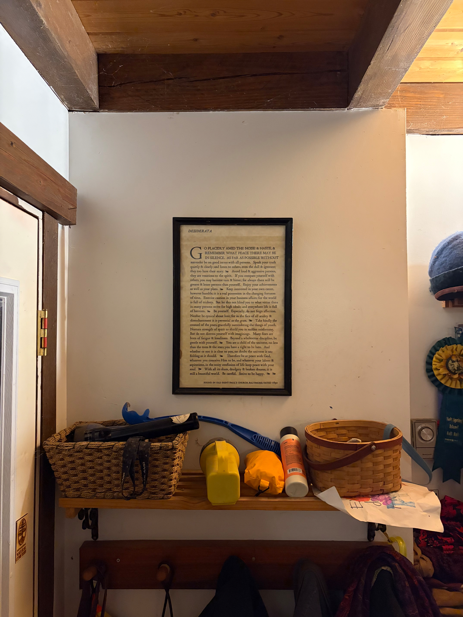 A small wooden shelf holds baskets and various items underneath a framed piece of text on a wall, with wooden ceiling beams above.