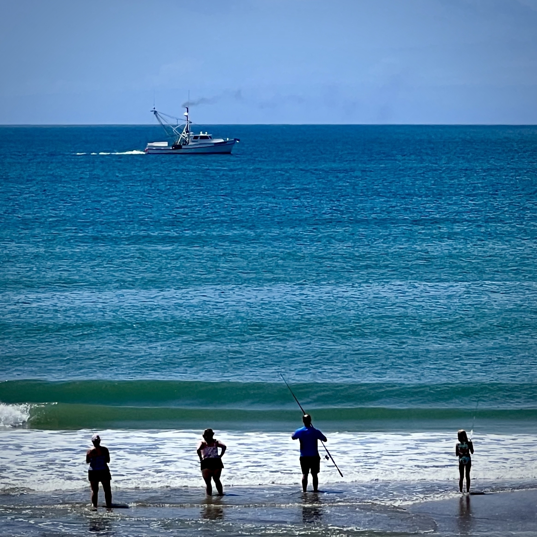 Distant image of four people standing in the surf of a beach. The water is blue and green with visible white foam. In the distance a fishing boat can be seen. 