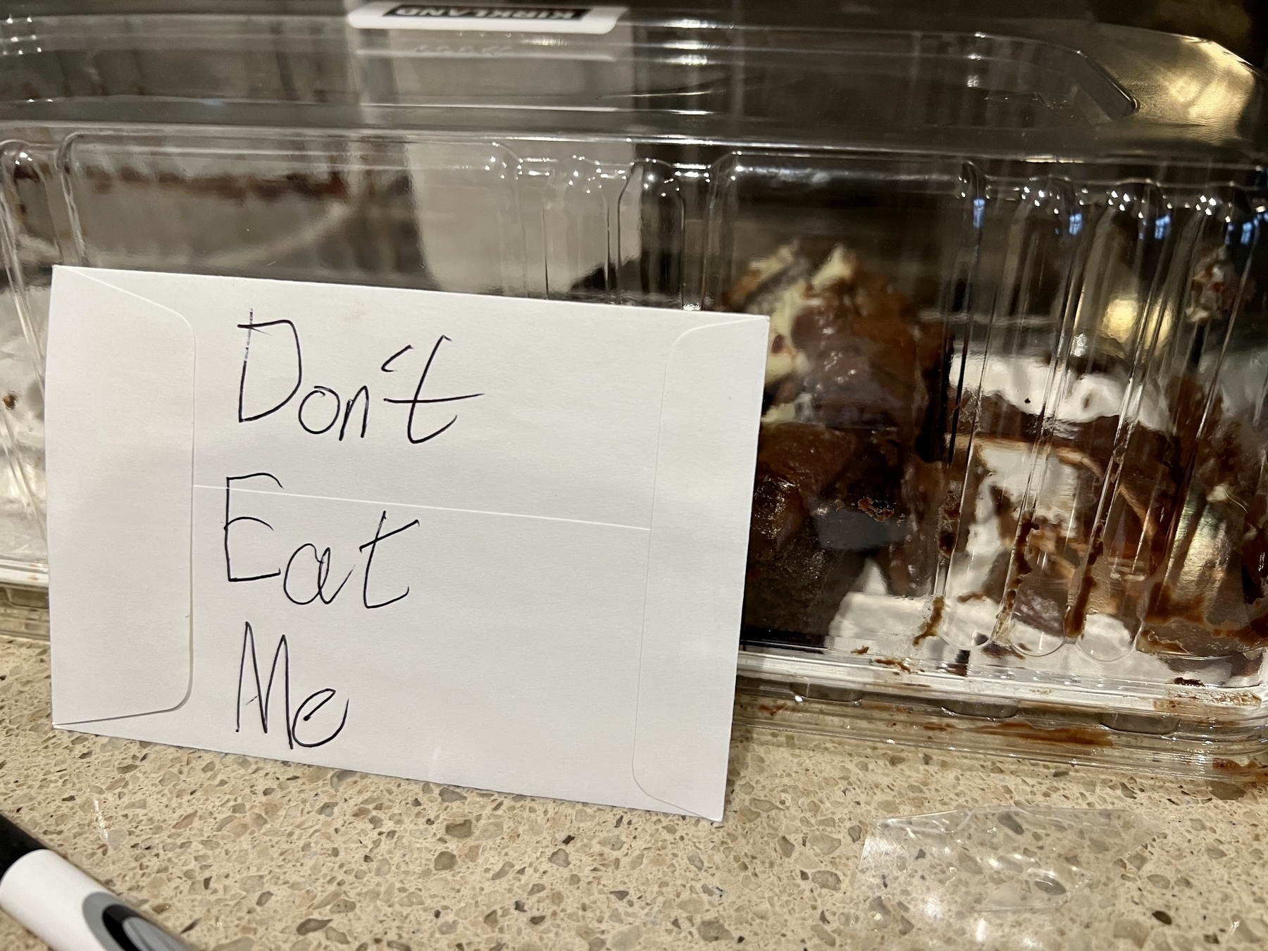 A sign reading “don’t eat me” in front of a piece of cake in a package on a kitchen counter. 