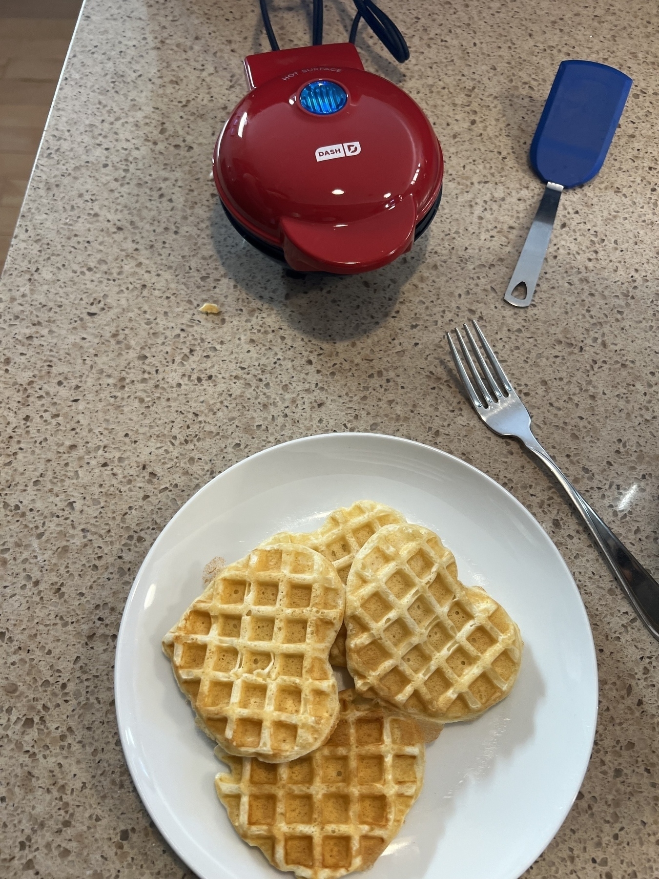 Heart shaped individual waffle maker with some waffles on a plate beside it. 