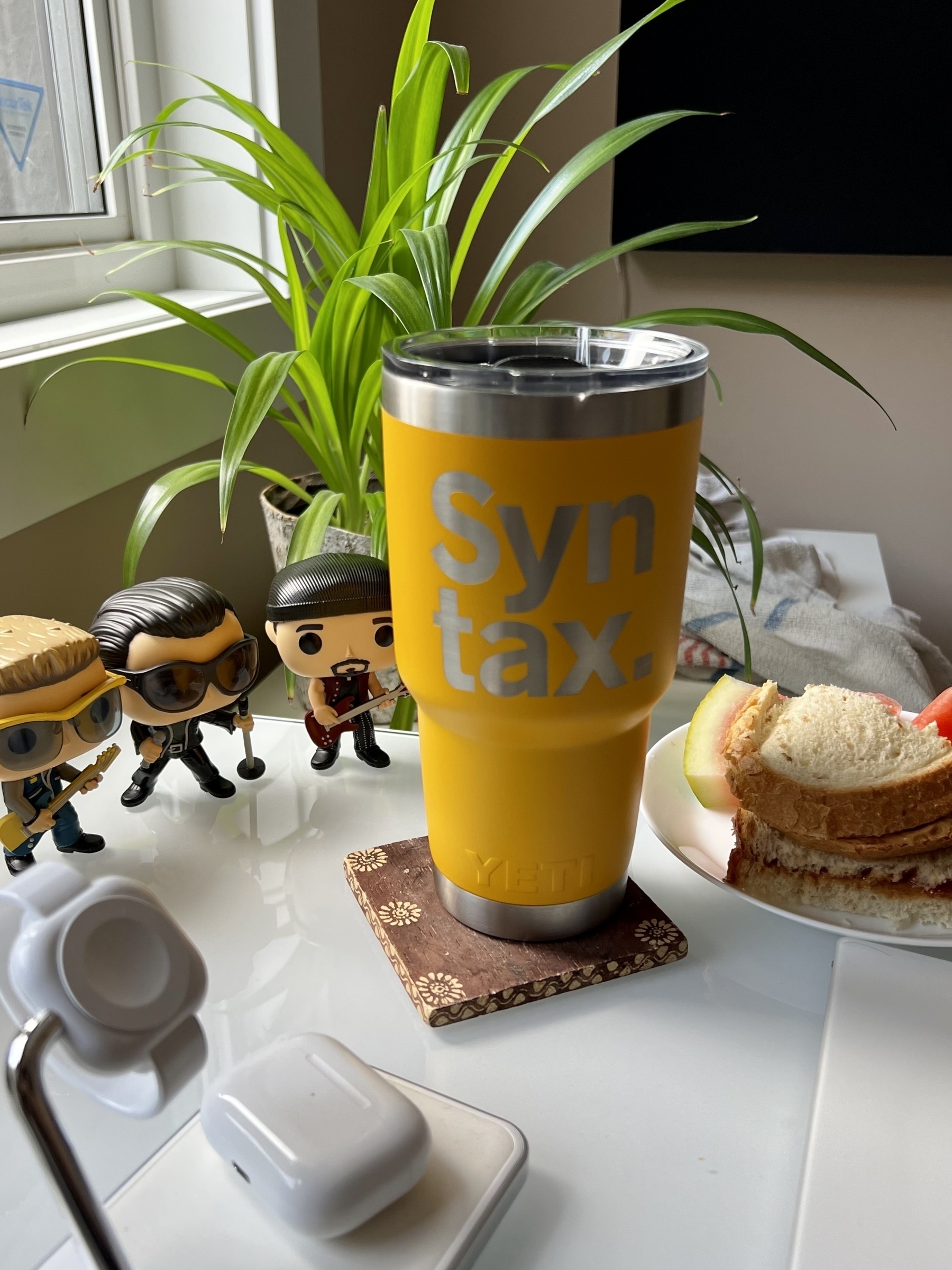 Yeti mug with Syntax.fm branding with U2 funkopop characters, a plant, and a sandwich beside it. 