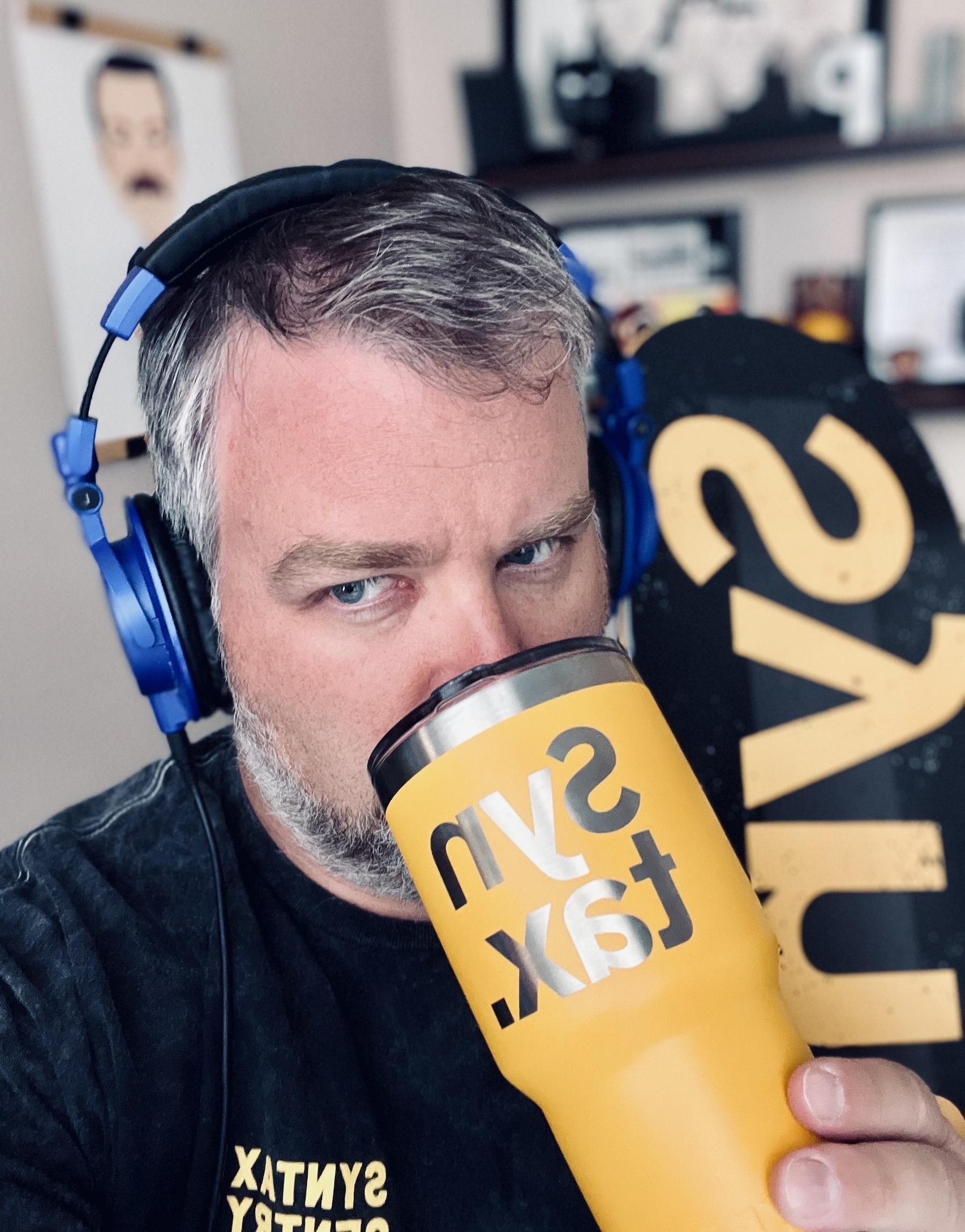 An adult male (me) holding a Yeti mug with Syntax branding, wearing headphones, holding a Syntax skate deck, and wearing a Syntax tshirt. 