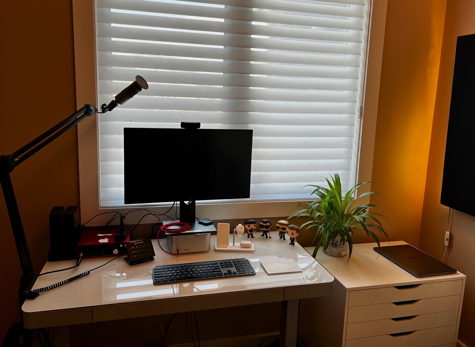 Photo of an office with a white desk, Dell monitor, Mac Studio, Heil PR40 mic on a boom arm, a white set of drawers, a yellow light glowing in the corner, and the members of U2 as funko pop toys on the desk.
