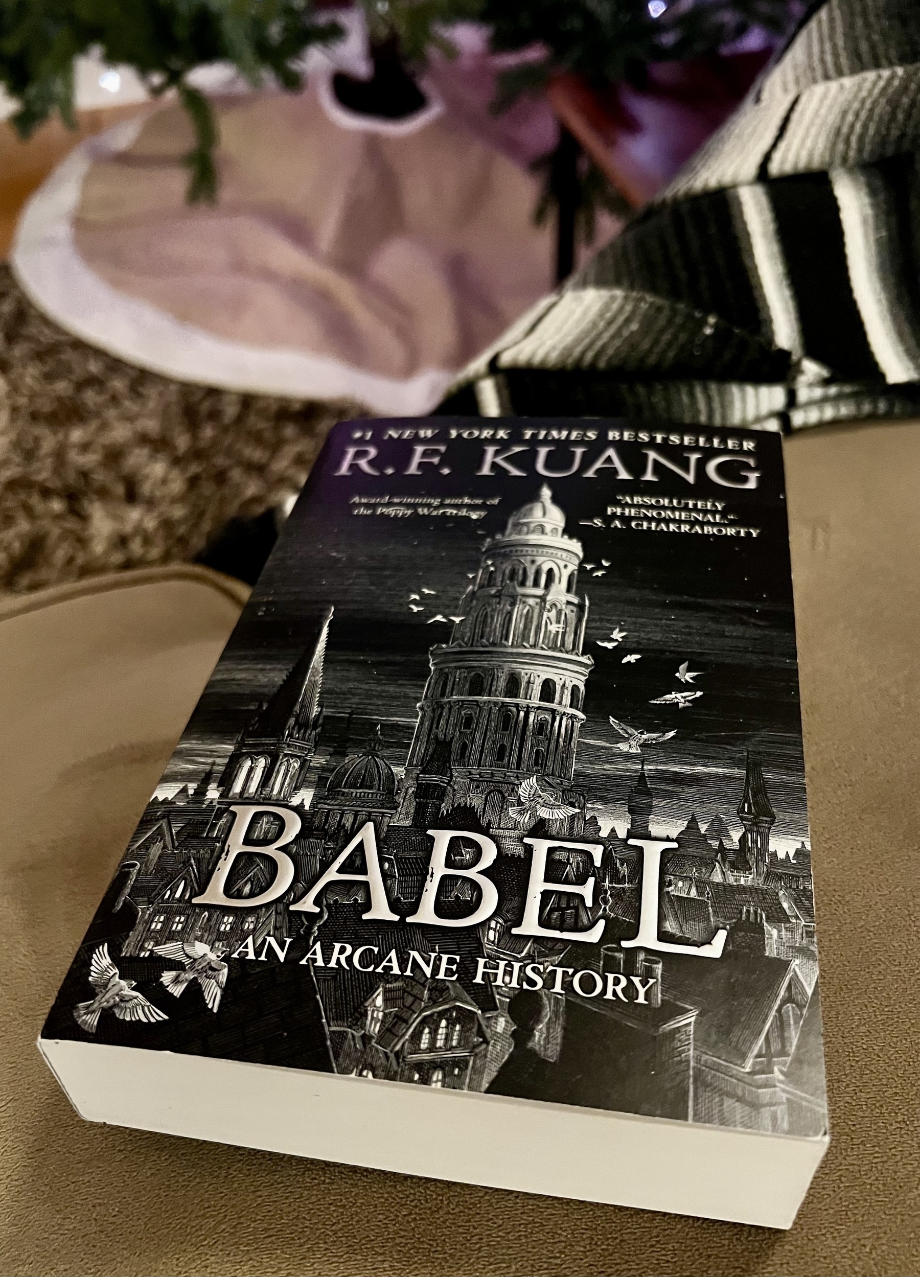 A paperback copy of "Babel: An Arcane History" by R.F. Kuang rests on a surface with a blurred background.