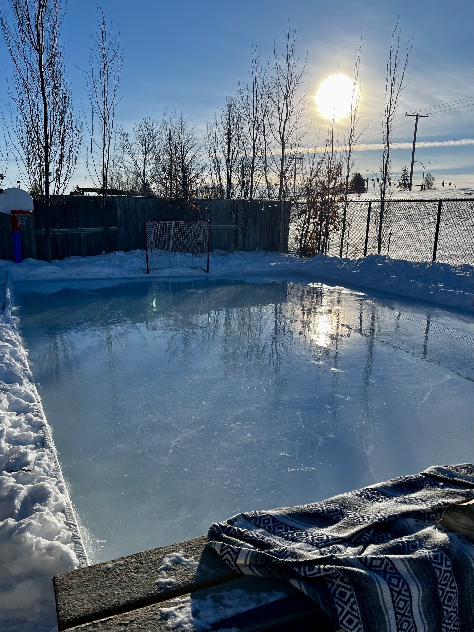 A backyard ice rink is surrounded by snow under a clear blue sky with the sun shining brightly.