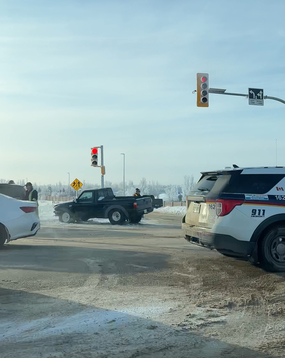 A small truck and a white car are in a snowy intersection near a red traffic light, with police and emergency responders present.