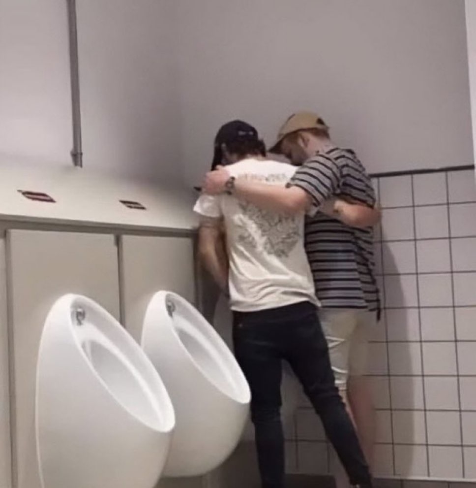 two dudes appear to be peeing into the same urinal together, arms around each other.