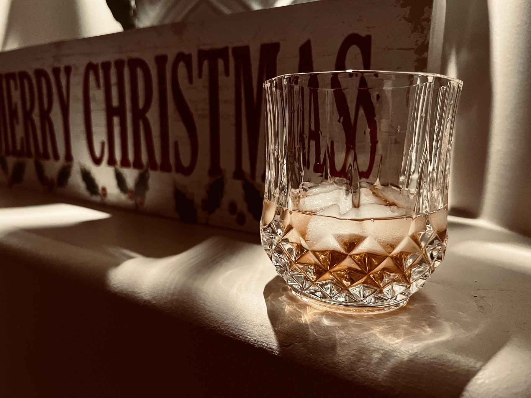 A glass with ice and whisky sits on a shelf next to a merry Christmas sign. 