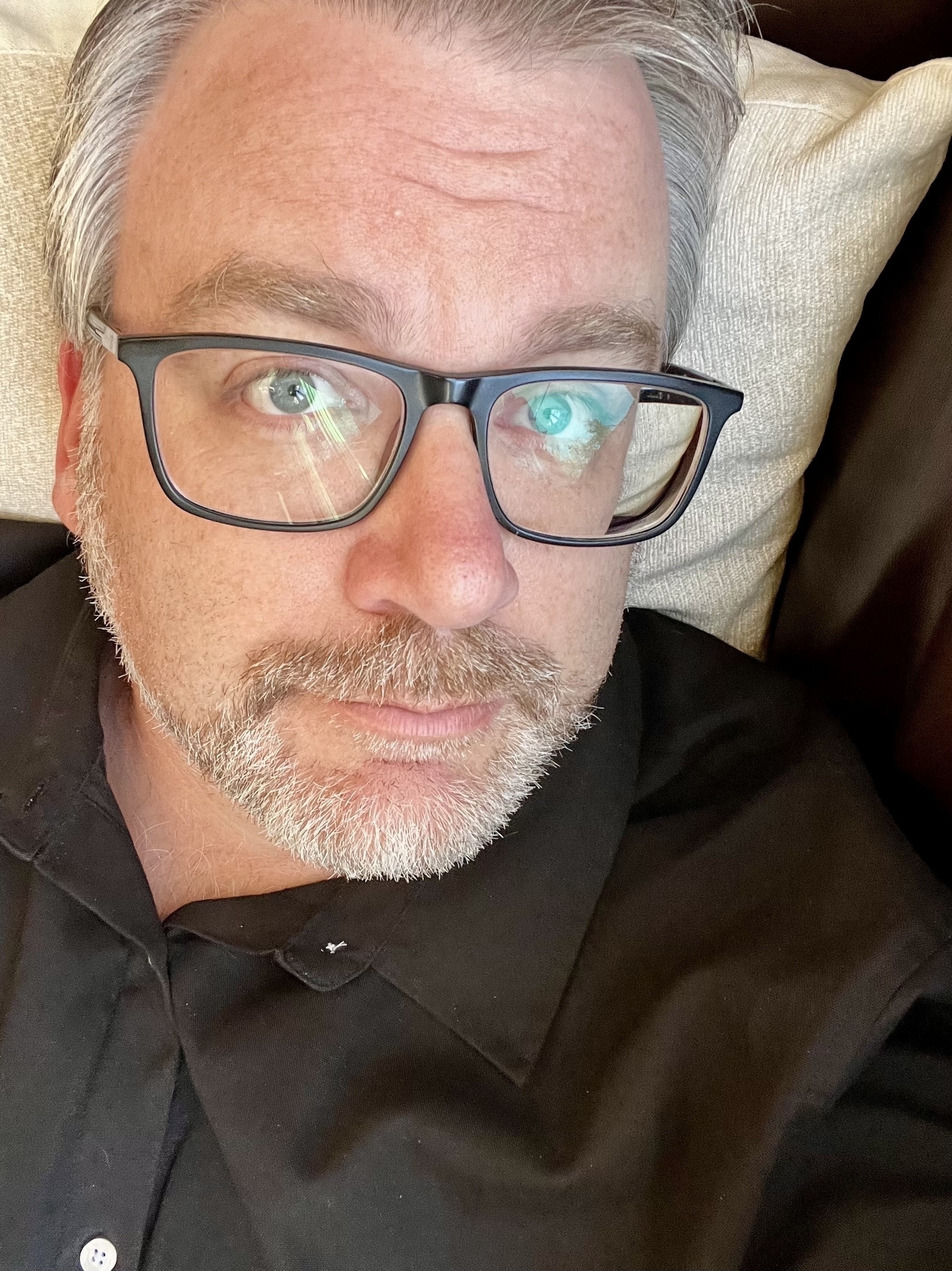 A man with glasses and a beard is lying down on a pillow, wearing a black shirt.