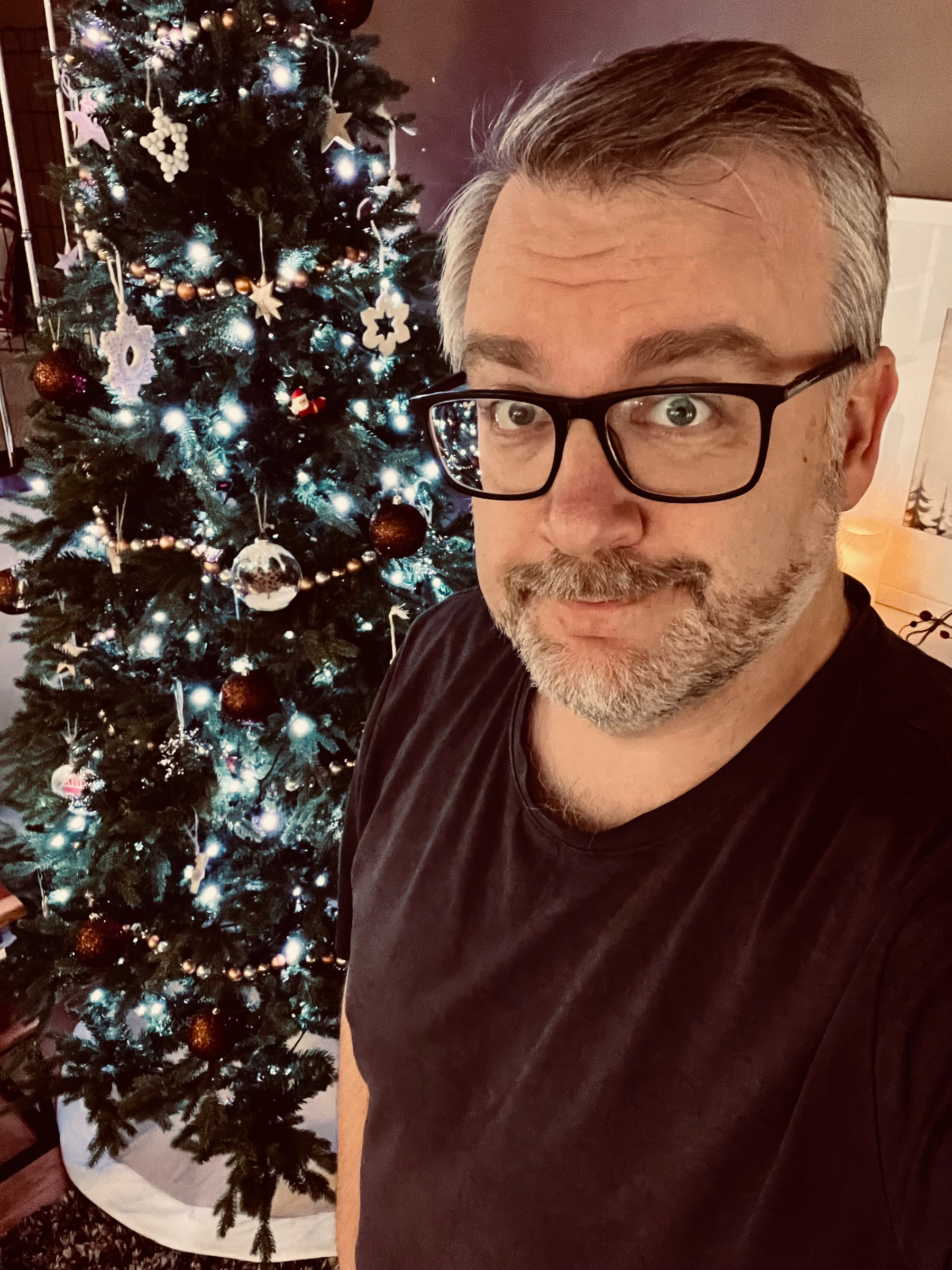 A person wearing glasses stands in front of a decorated Christmas tree.
