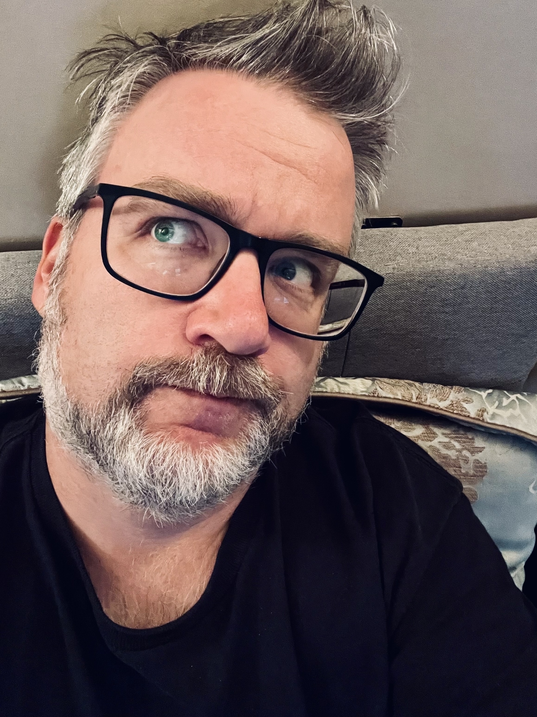 A bearded individual with glasses looks off to the side, with a thoughtful expression, seated against a cushion.