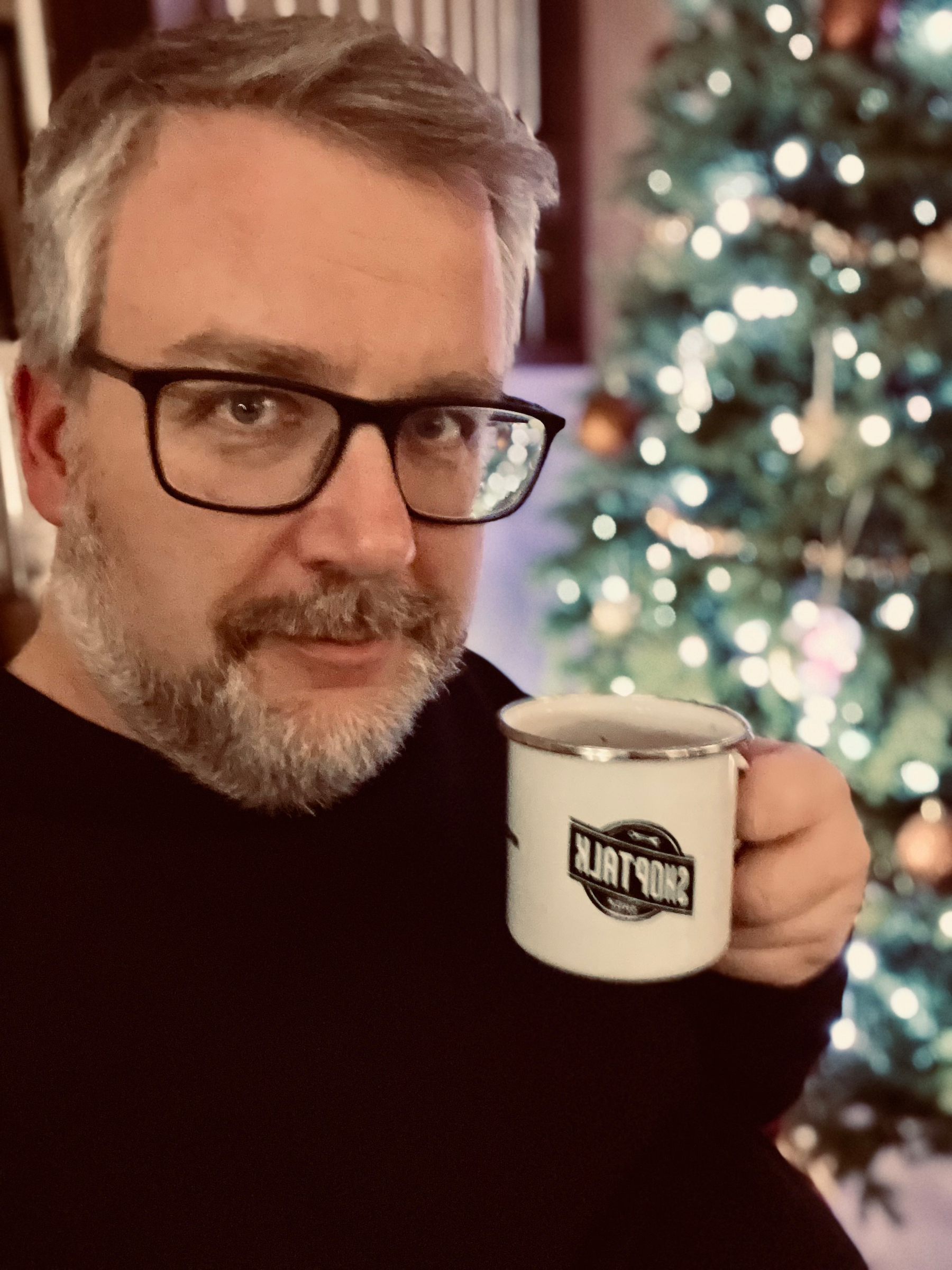 A person holds a mug while standing in front of a decorated Christmas tree with lights. The mug is a Shoptalk Show mug. 