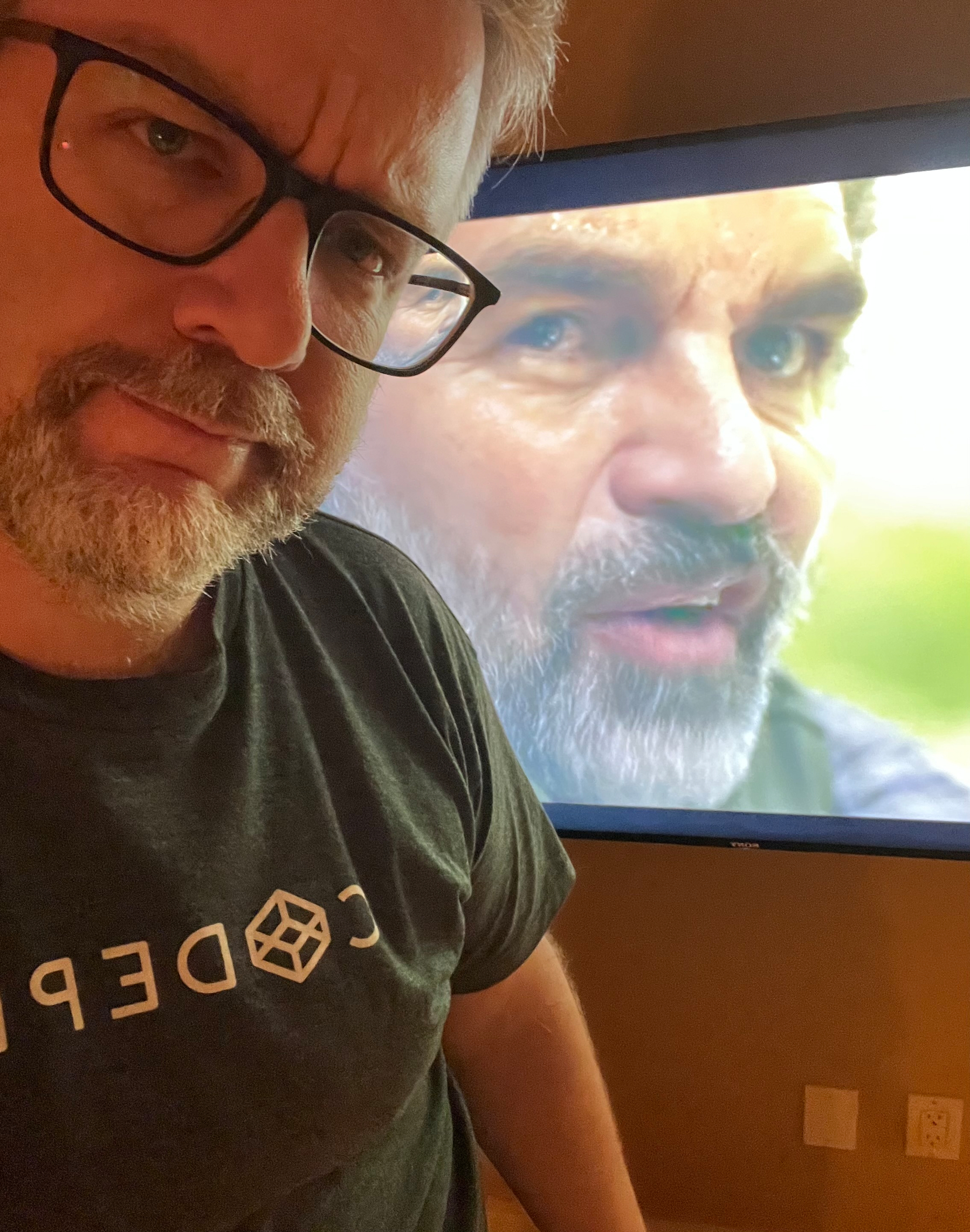 A person wearing glasses and a CodePen t-shirt stands with a TV screen in the background displaying a close-up of another person's face, Mark Ruffalo the actor. 