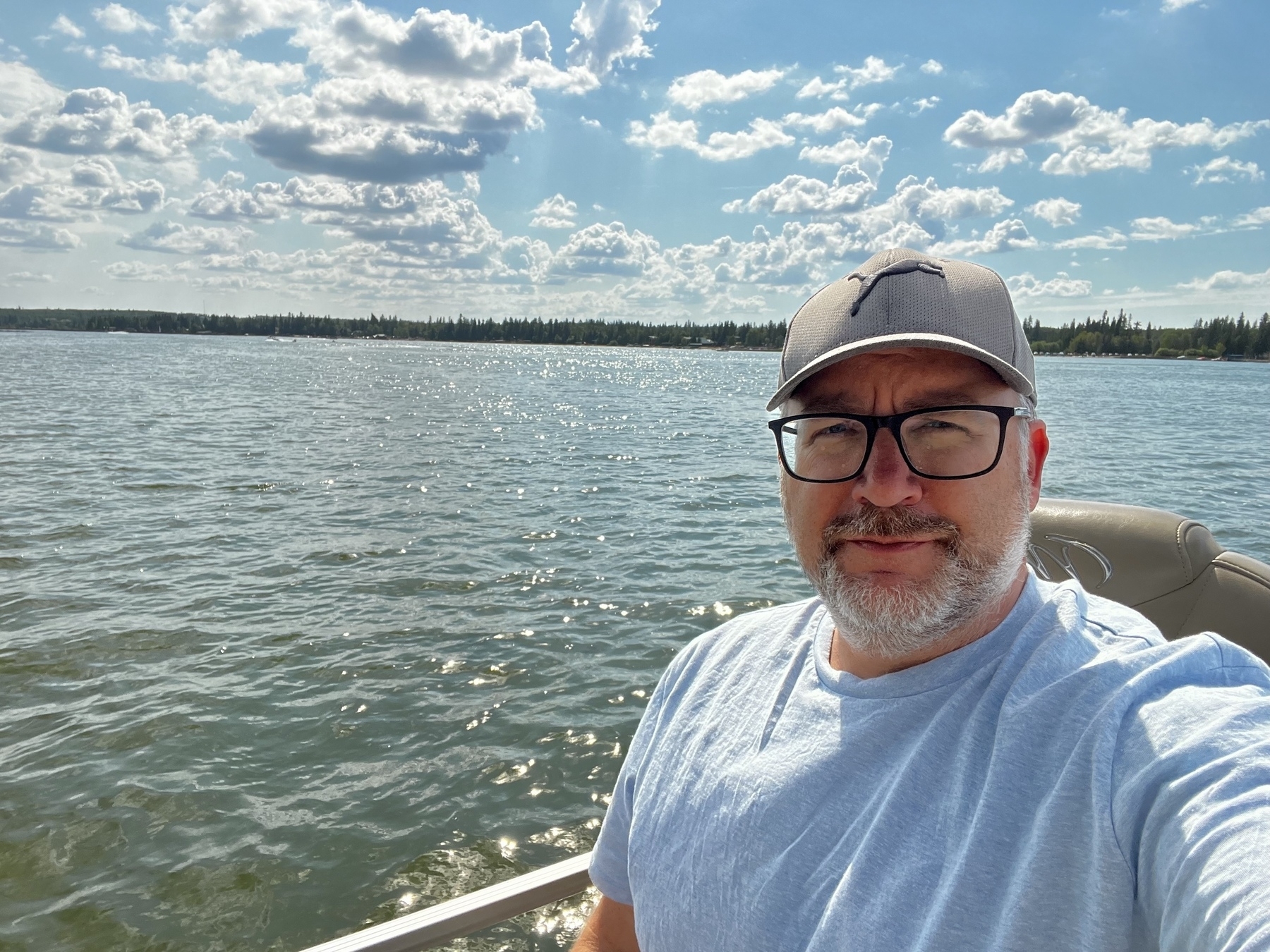 A man wearing glasses and a cap is sitting on a boat with a vast body of water and a cloudy sky in the background.