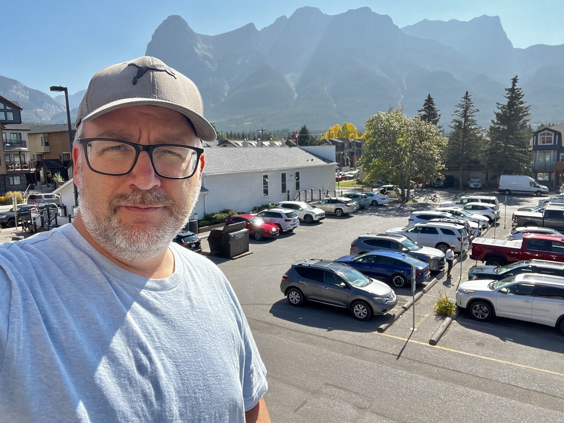 A man wearing glasses and a cap is taking a selfie with a parking lot and mountains in the background.