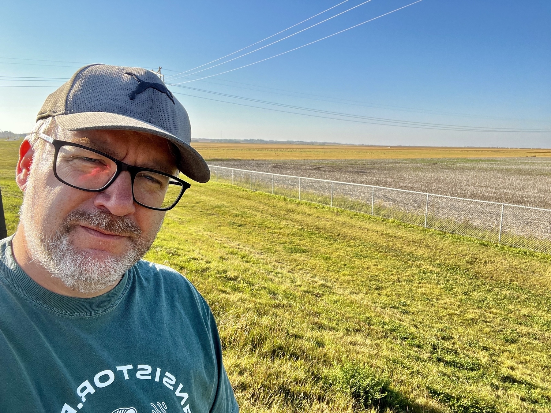 Selfie of a man with a hat, glasses, and a beard staring in front of a prairie field with a blue sky behind. 