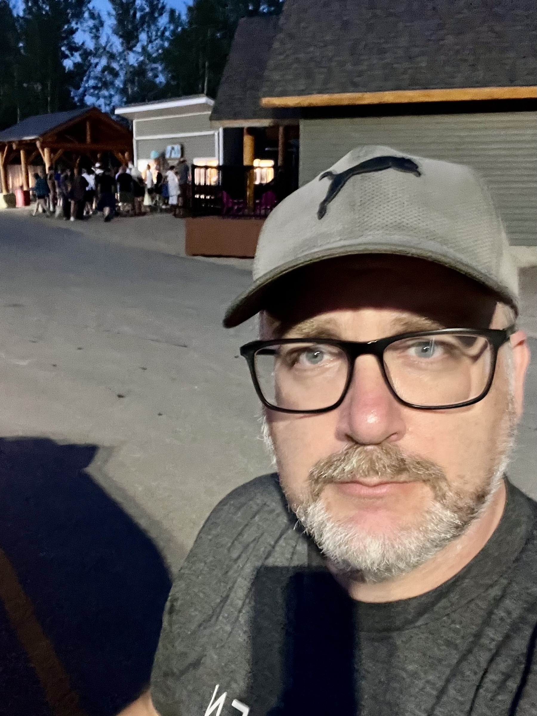 A man wearing glasses and a cap takes a selfie in front of a building with a group of people gathered near it.
