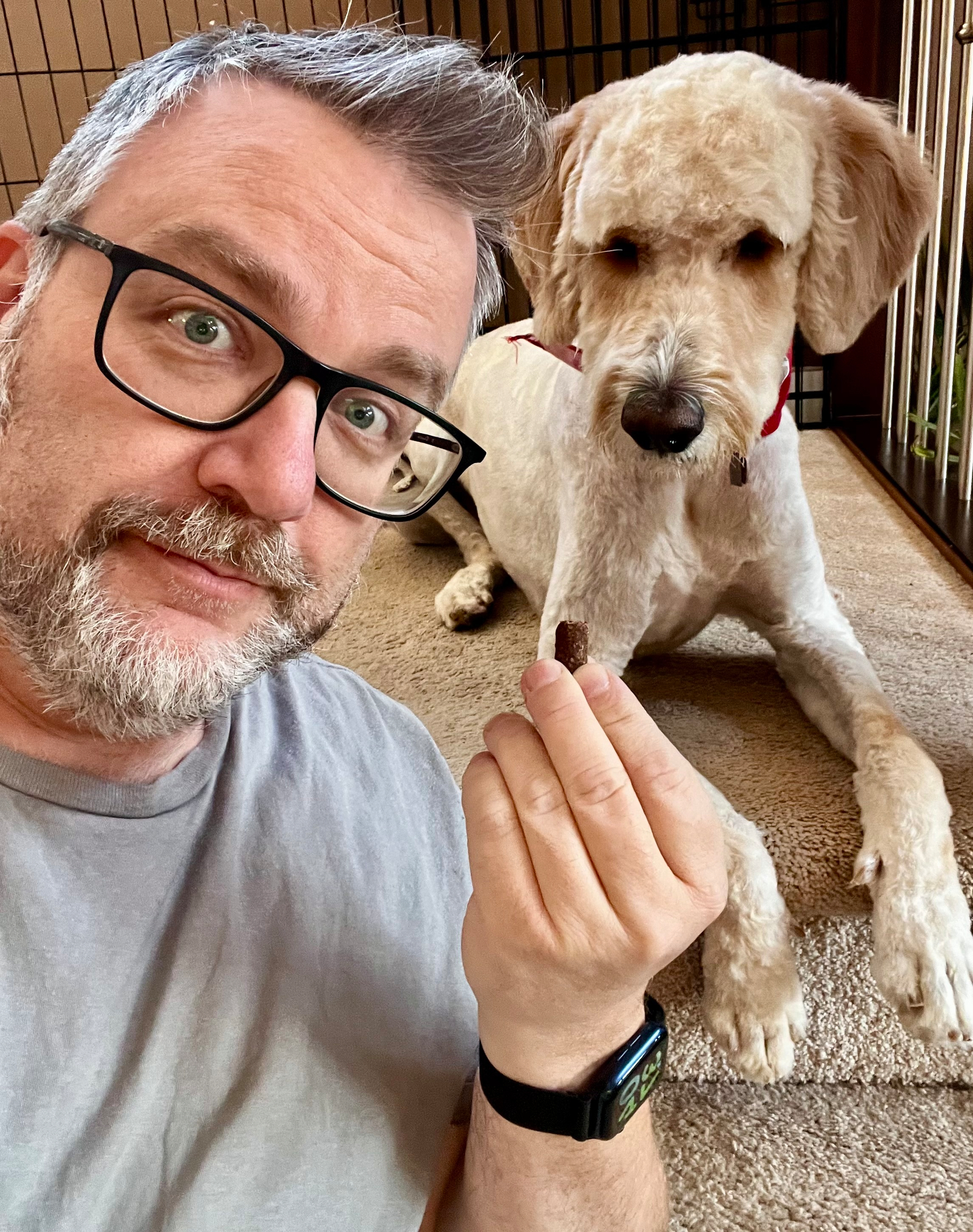 A man with glasses holding a treat poses with a dog lying down nearby.