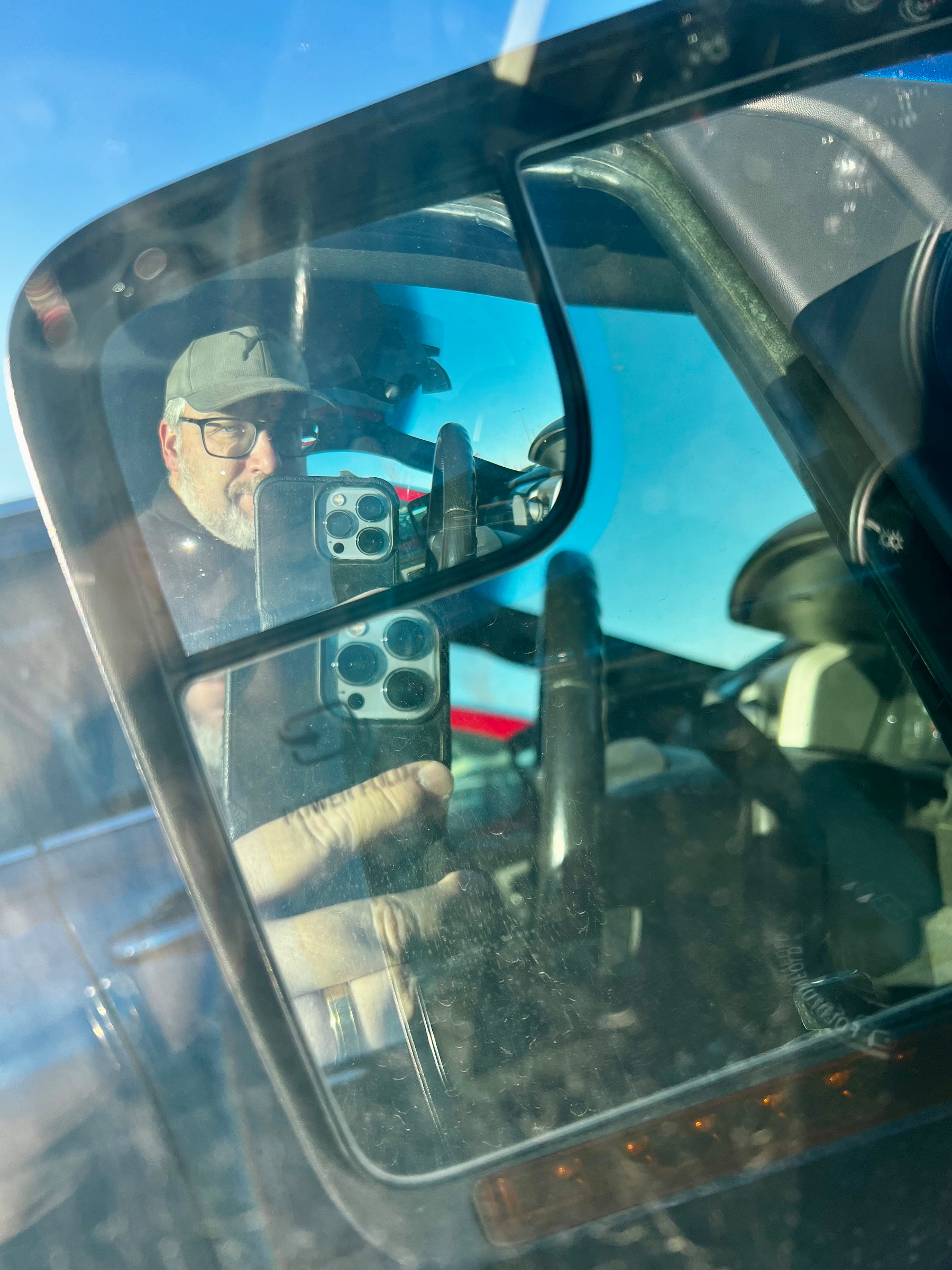 A reflection in a side mirror shows a person holding a smartphone, with part of a vehicle and blue sky visible.