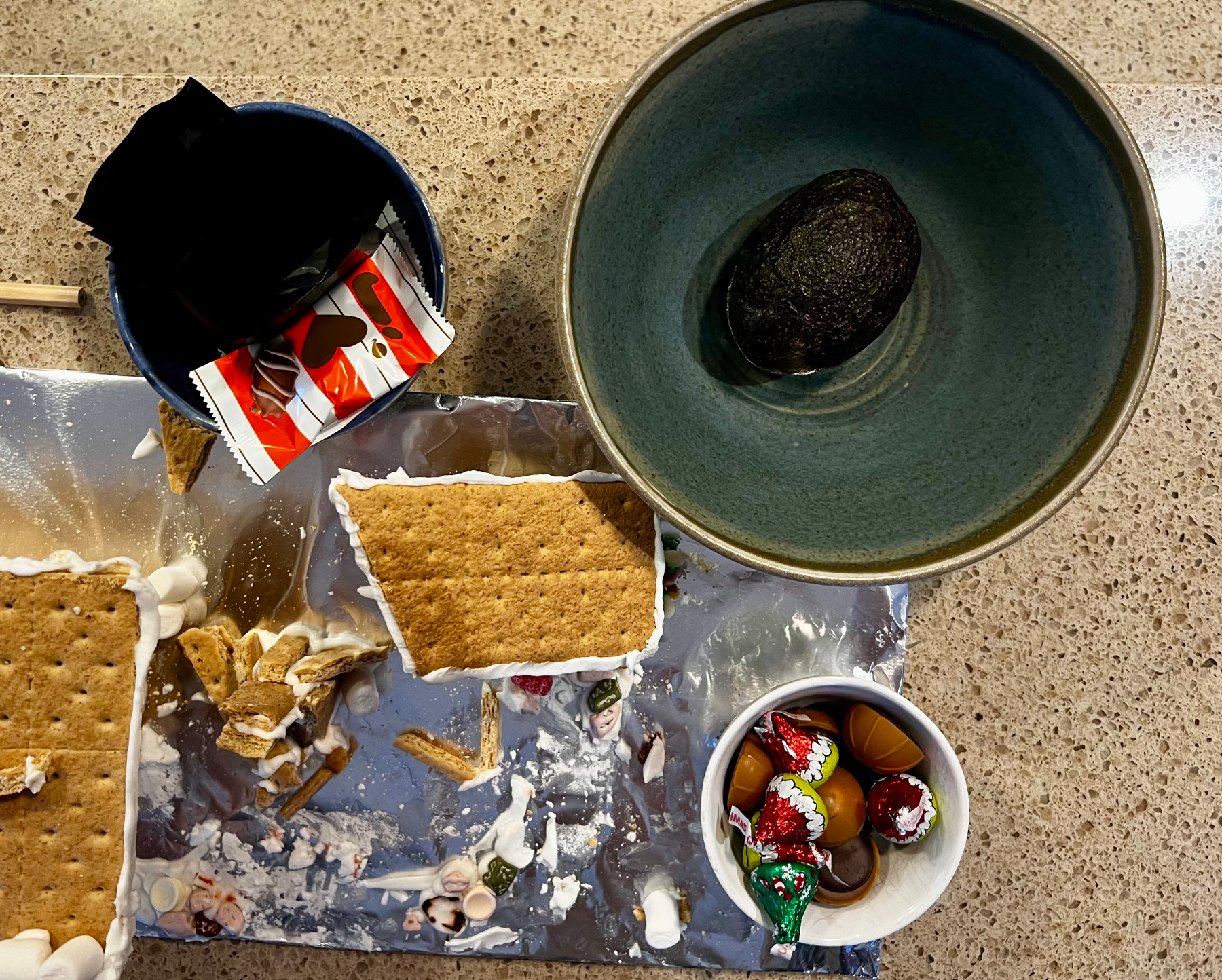 A messy tabletop is covered with broken graham crackers, marshmallow remnants, a bowl with an avocado, and a small bowl containing assorted chocolates.