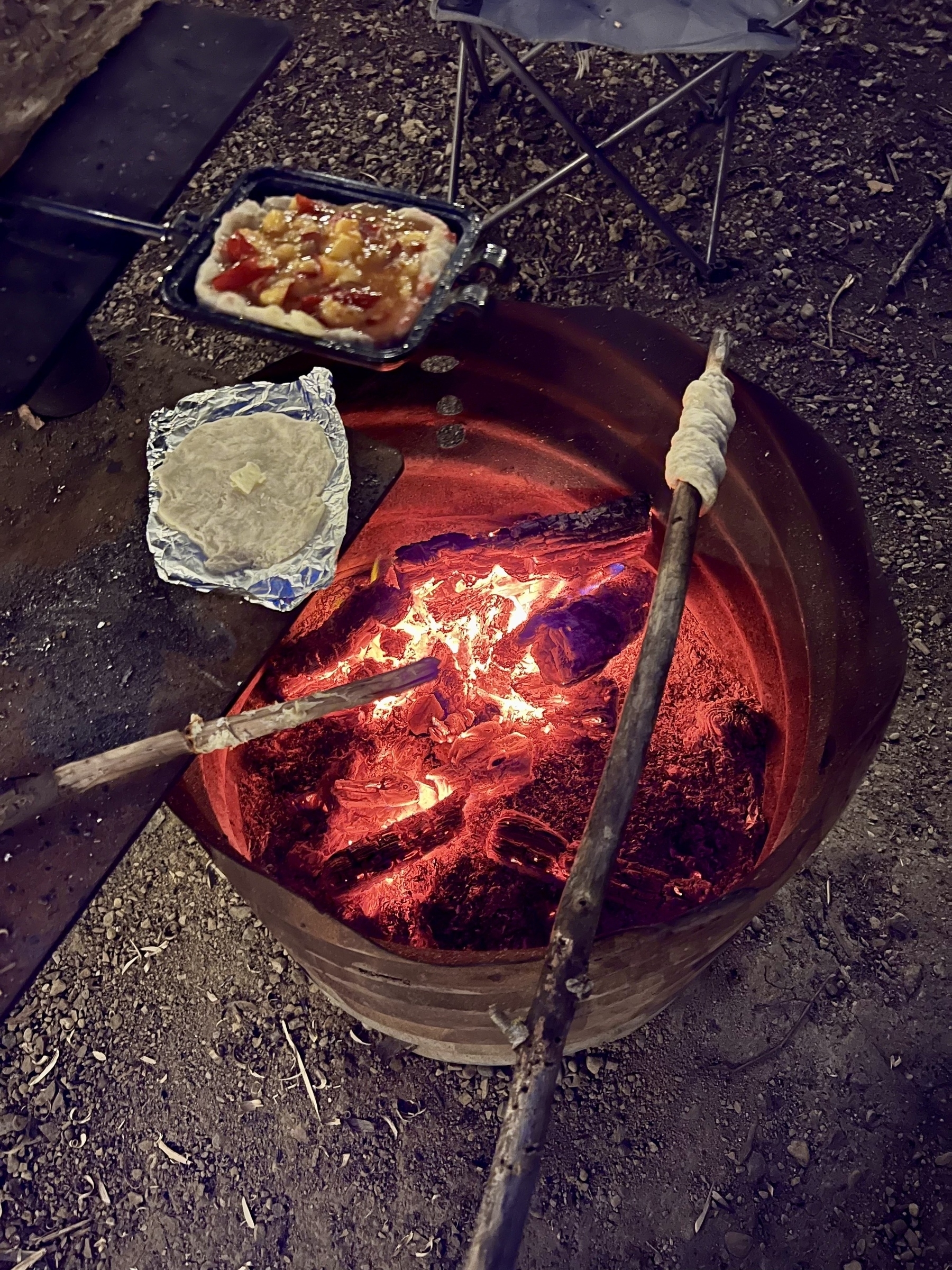 Cooking over a campfire includes a bread-dough-wrapped stick and a pan of pizza or similar food resting on embers inside a barrel.