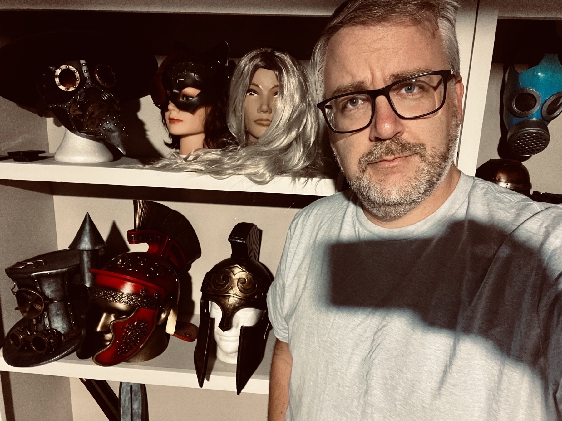 A person with glasses and facial hair stands in front of shelves displaying various helmets and masks.