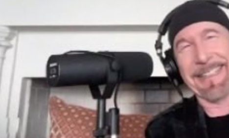 A screenshot of a Zoom call showing U2’s The Edge smiling in a room with headphones on and a Shure SM7B microphone in front of him.