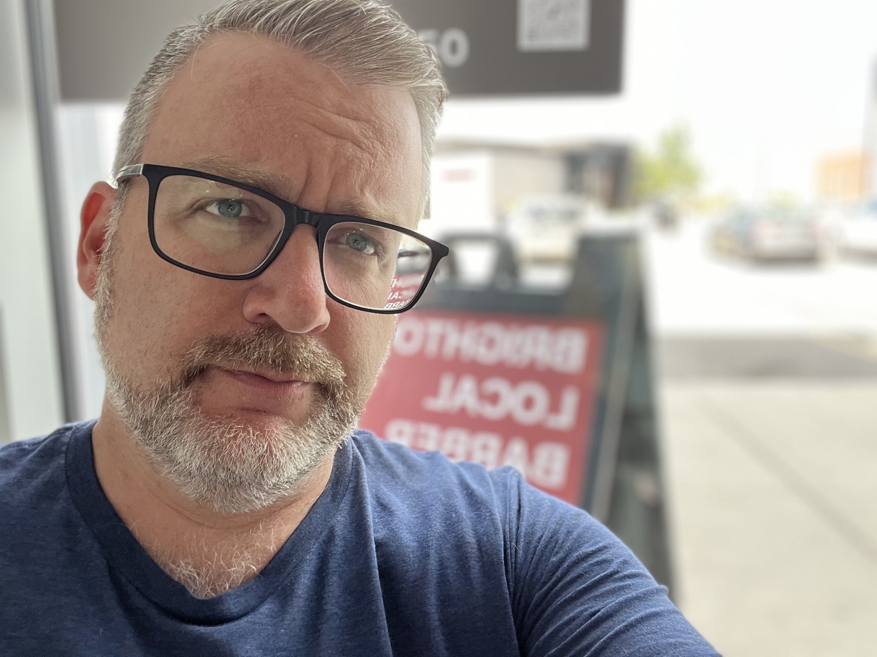 A man with glasses and a beard is taking a selfie in front of a window, with a blurred sign in the background.