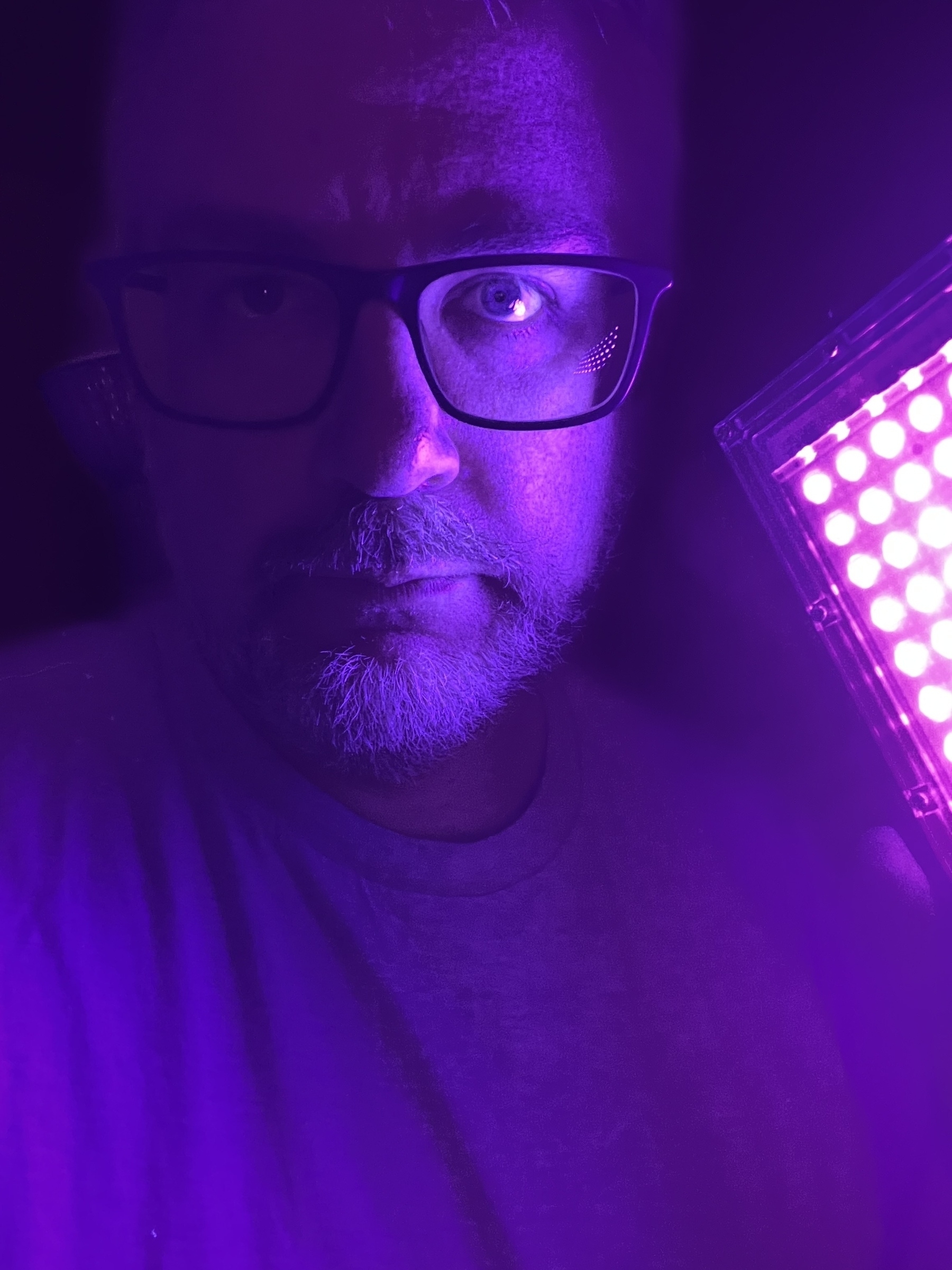 A person is illuminated with purple light from an LED panel, wearing glasses and a neutral expression.