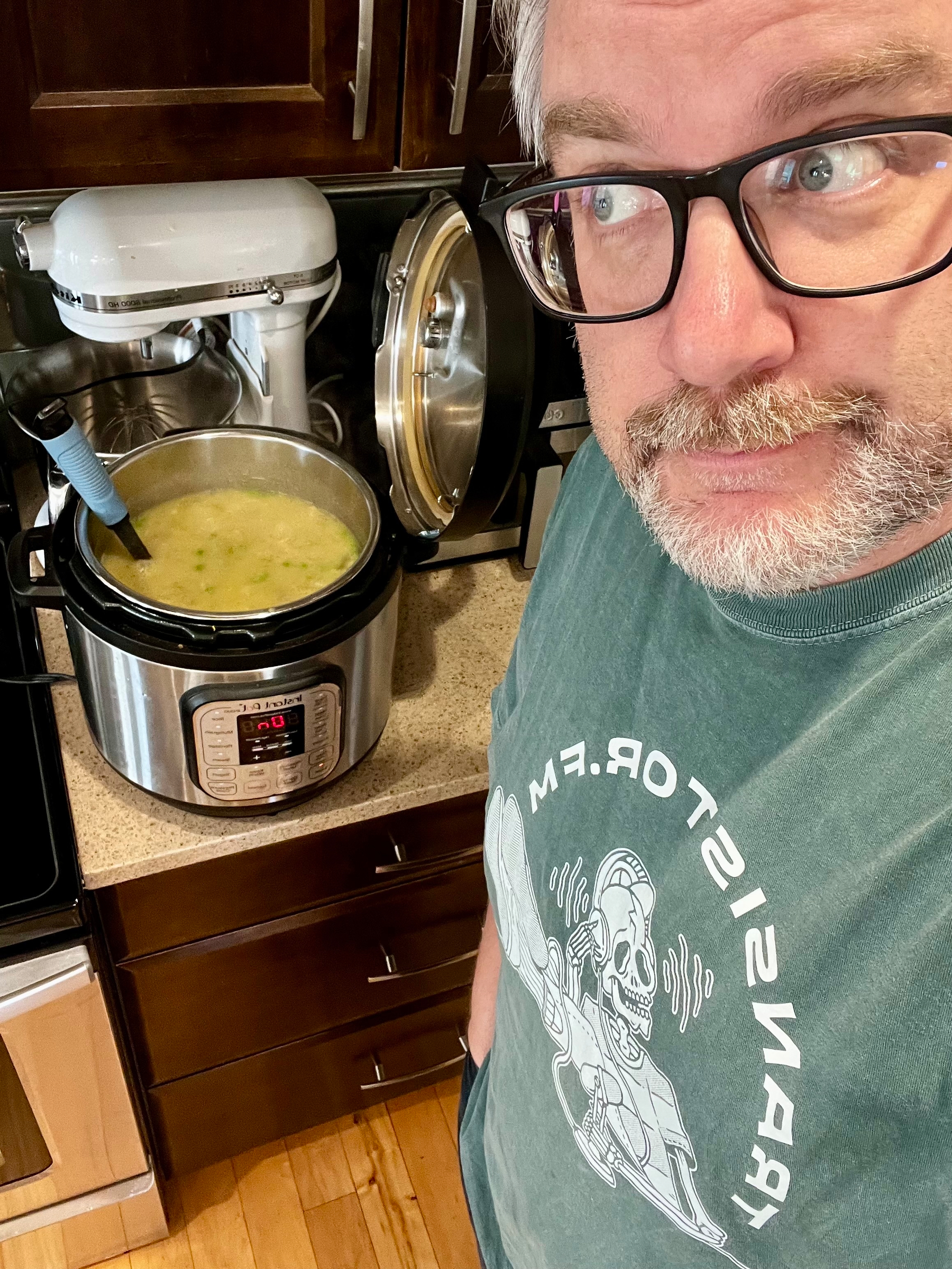 A person wearing glasses and a graphic T-shirt stands in a kitchen next to an Instant Pot filled with soup.