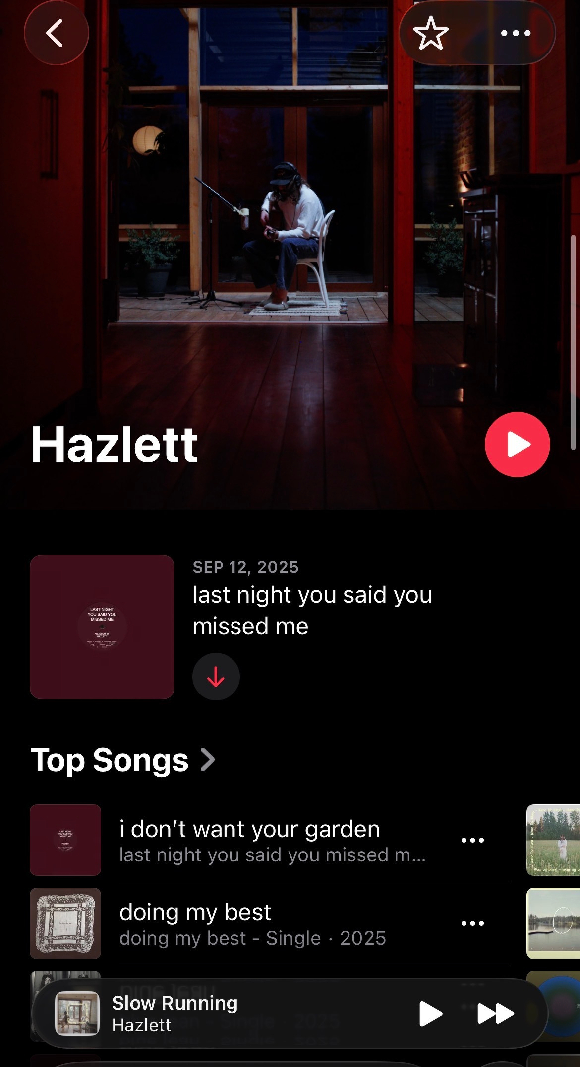 A person is sitting and playing guitar inside a dimly lit room, as shown on a music streaming app screen featuring Hazlett's songs.