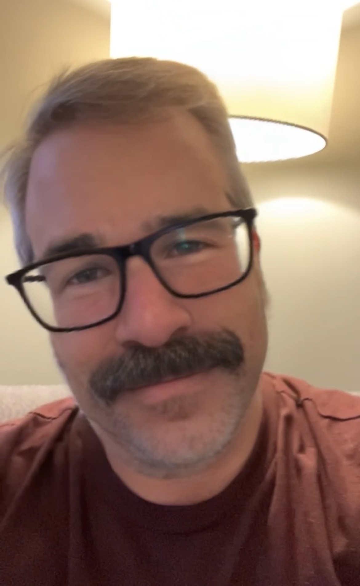 A person with glasses, a mustache, and a maroon shirt is sitting in front of a brightly lit lamp.