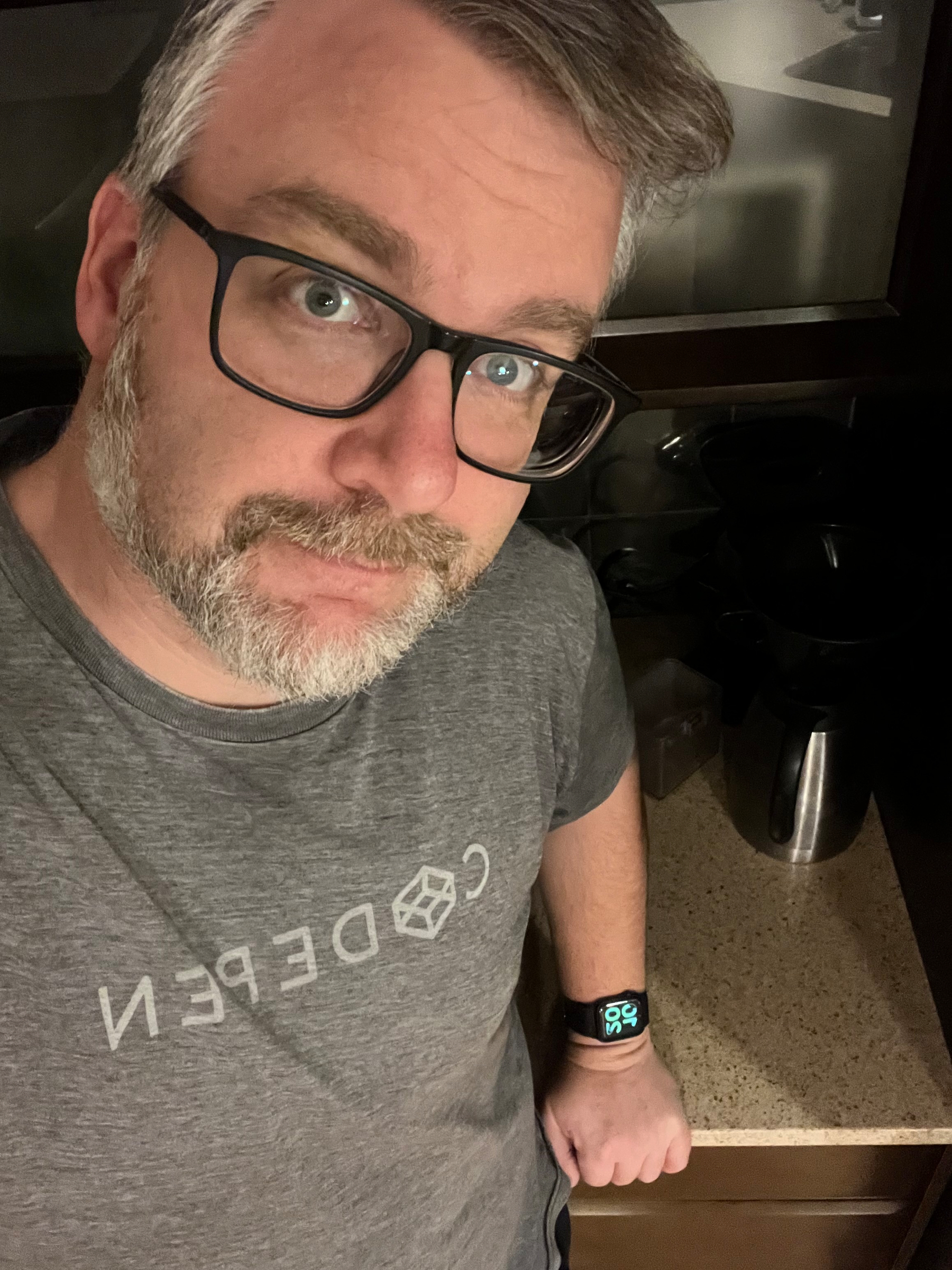 A person with glasses and a beard is wearing a gray t-shirt and a smartwatch, standing in a kitchen.