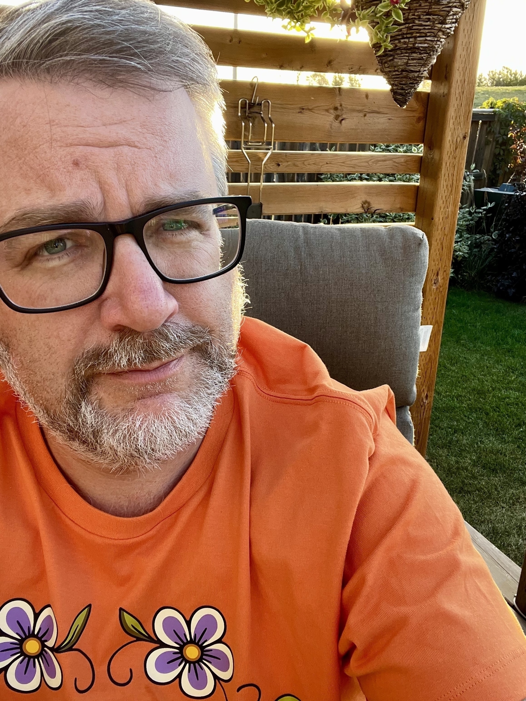A man in glasses and an orange shirt with floral designs is sitting outdoors by a wooden fence.