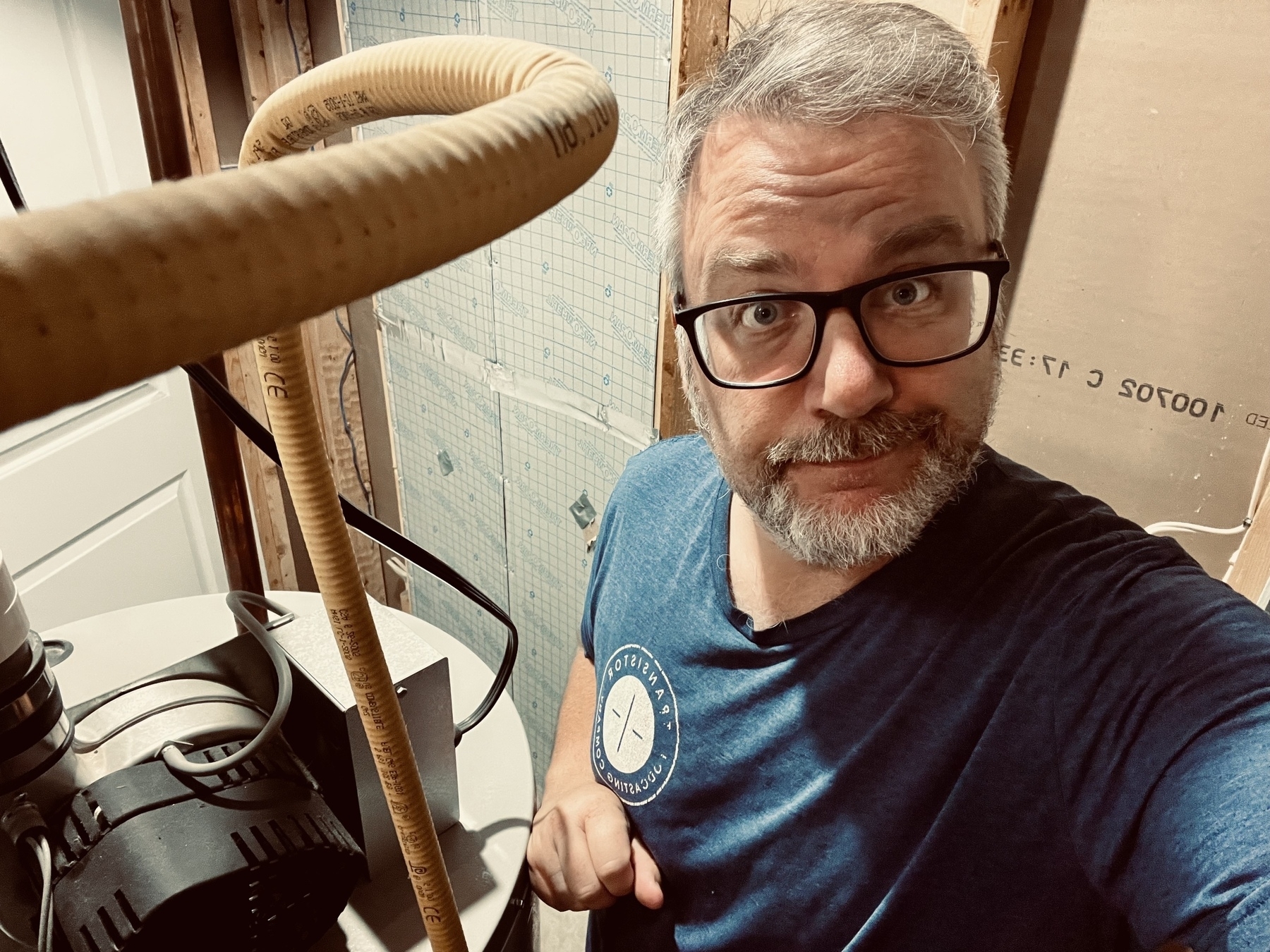 A person with glasses and a beard is standing in a room with industrial equipment and pipes.
