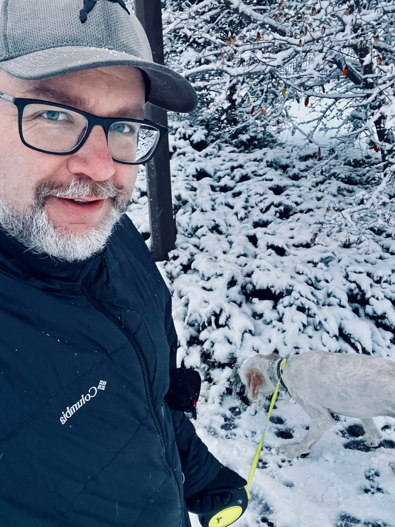 A man in winter clothing takes a selfie while walking a dog in a snowy environment.