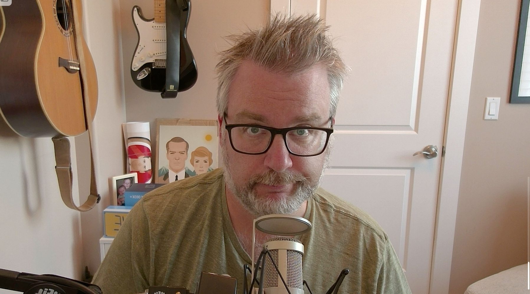 A man with glasses and a beard sits in front of a microphone, surrounded by musical instruments and artwork on the wall.