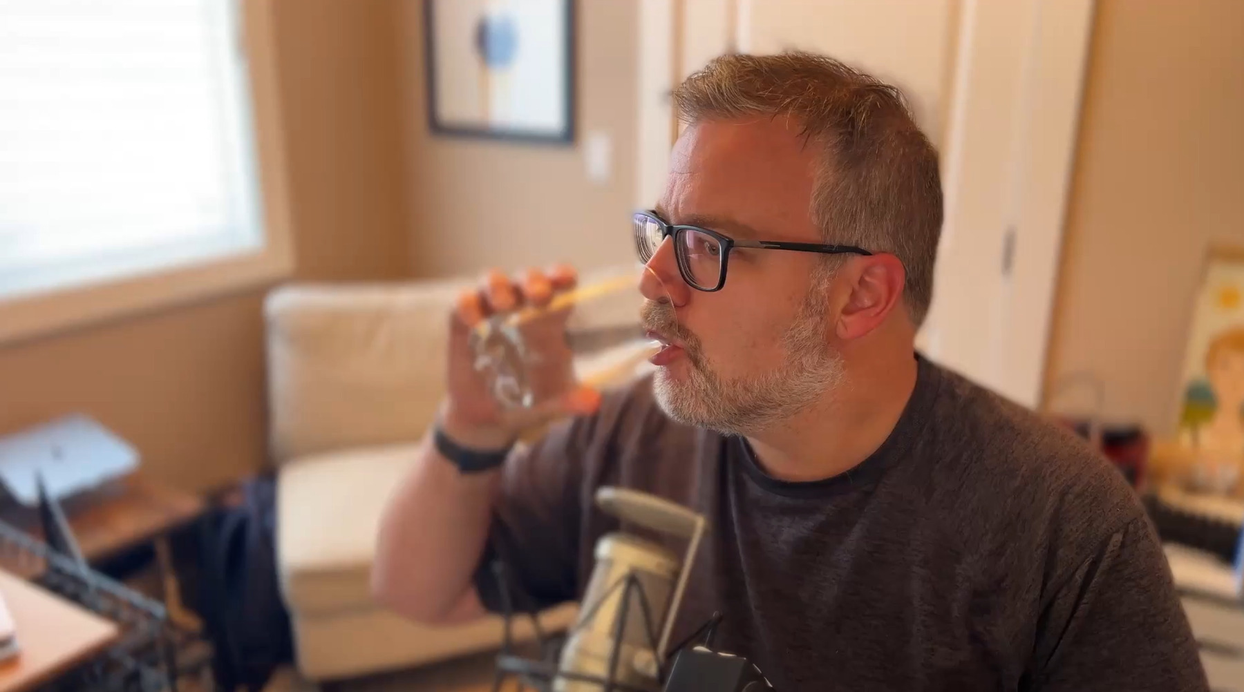 A man with glasses is drinking from a glass in a cozy room with a microphone nearby.