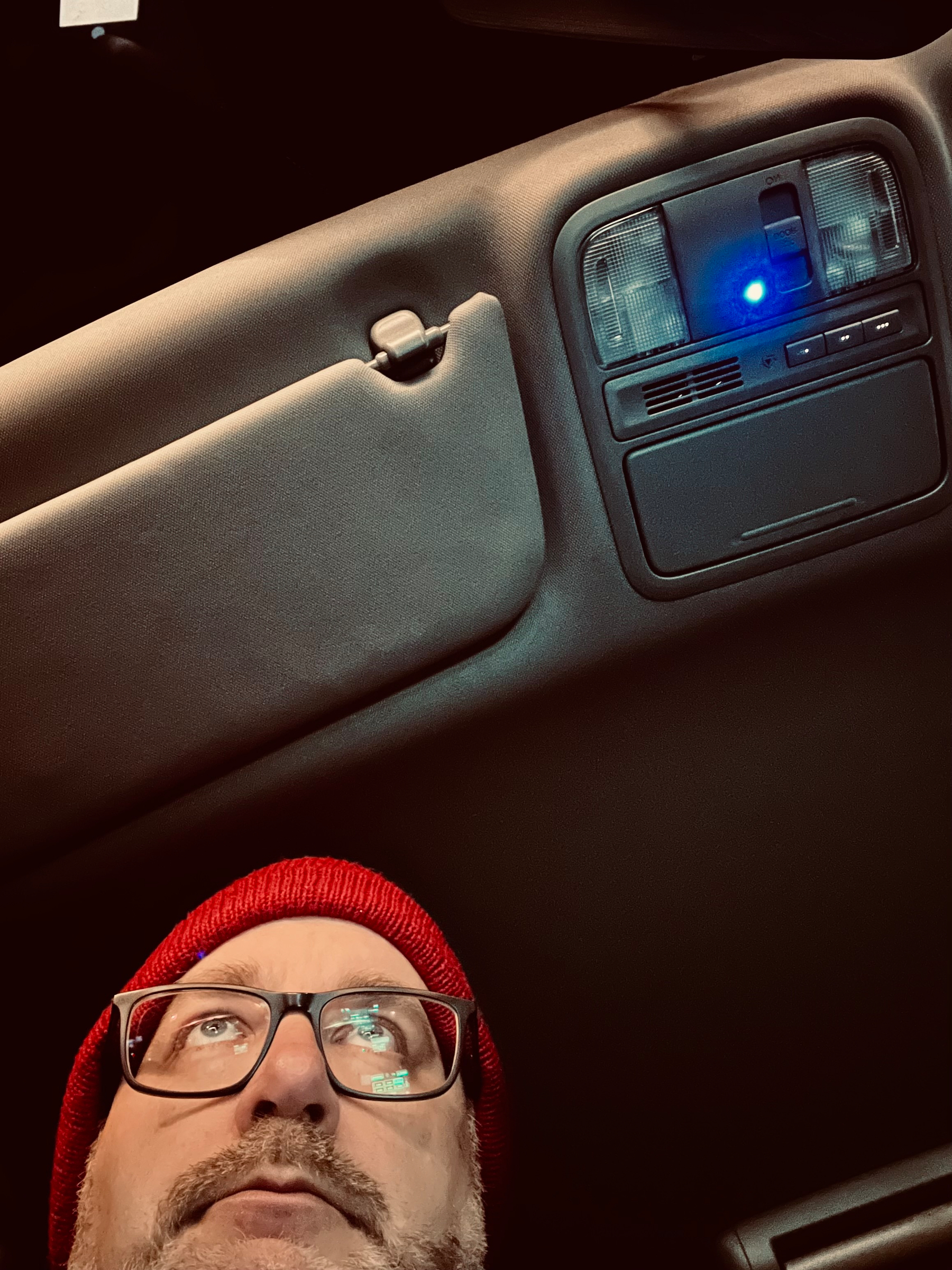 A person wearing glasses and a red beanie is sitting inside a car, looking up at the car's interior light panel on the ceiling.