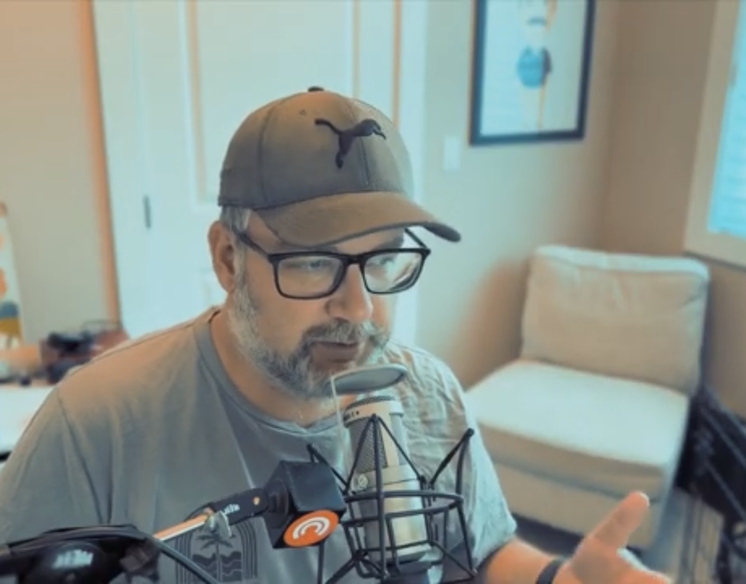 A man wearing a cap and glasses is speaking into a microphone in a room with a chair and wall art.