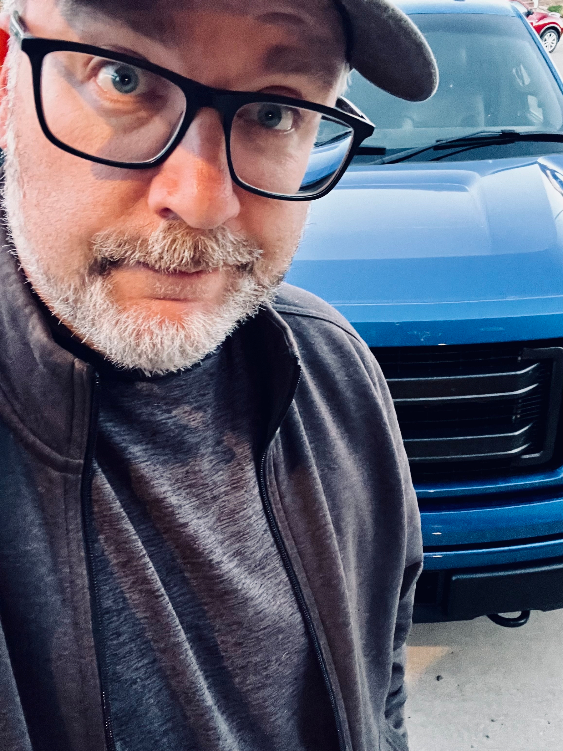 A person wearing glasses and a cap stands in front of a blue vehicle.