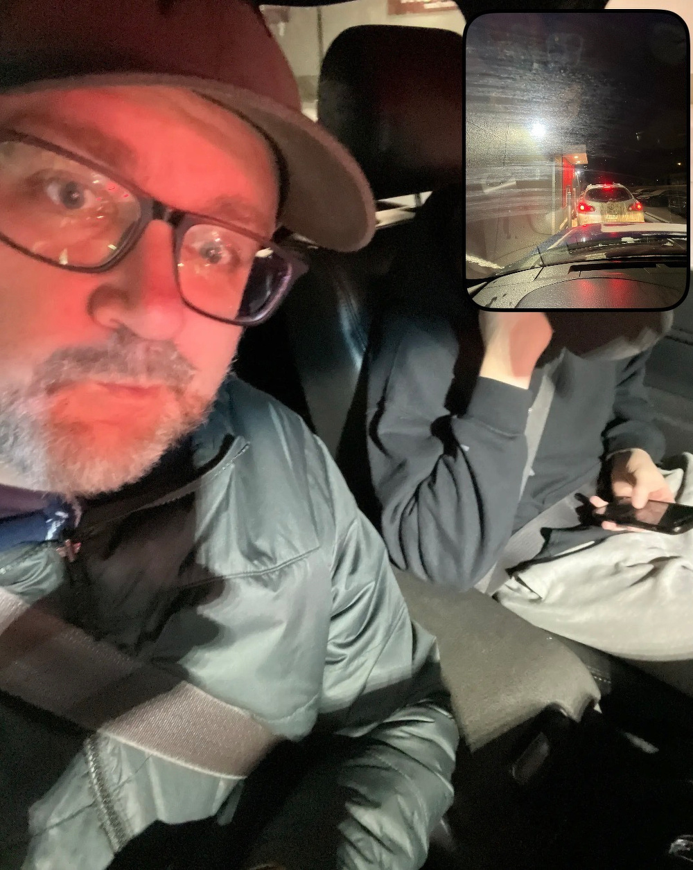 Two people are sitting in a car, with a red light illuminating the scene and a view of traffic outside through the windshield.
