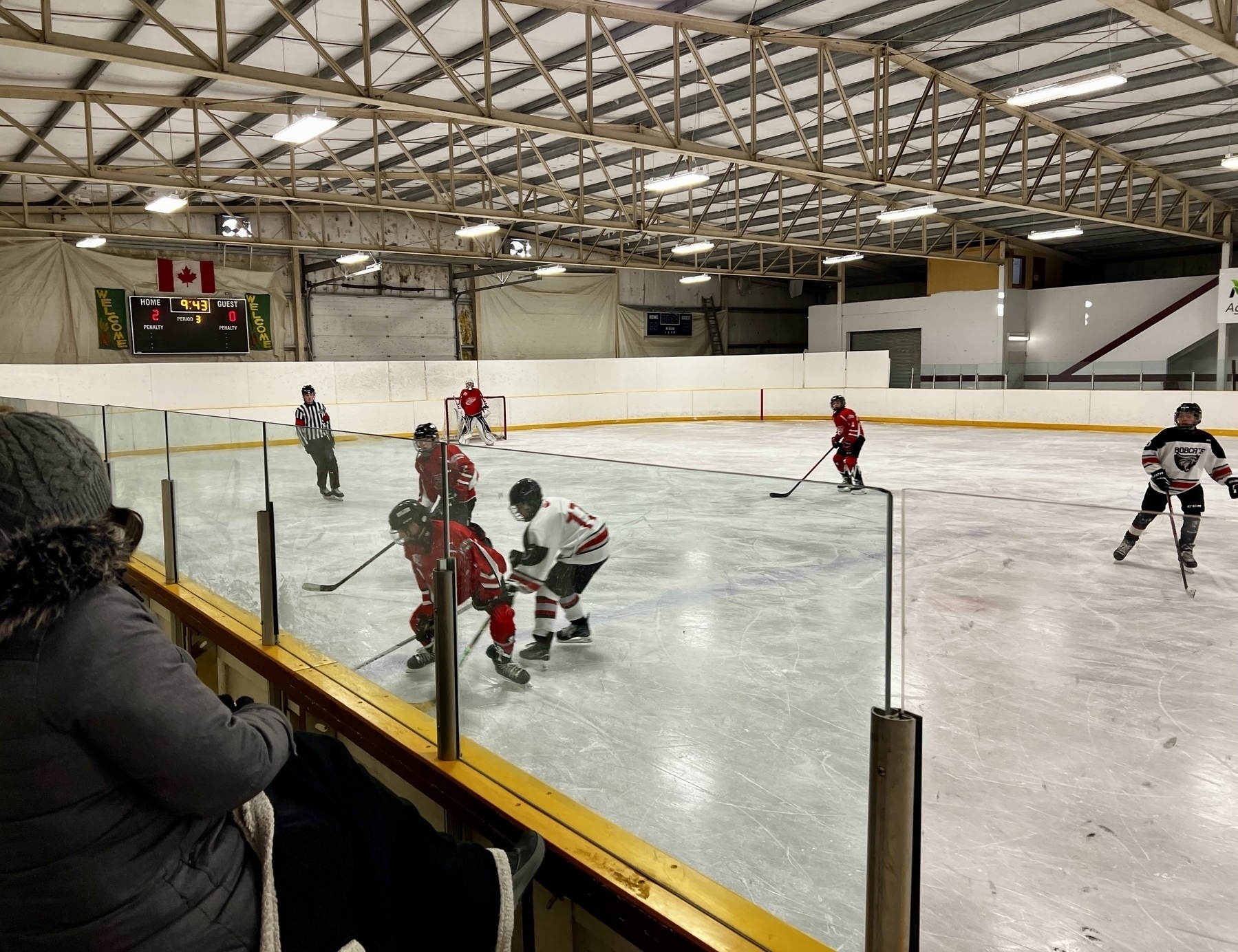 Hockey players on an indoor ice rink are engaged in a game while a spectator watches from behind a glass barrier.