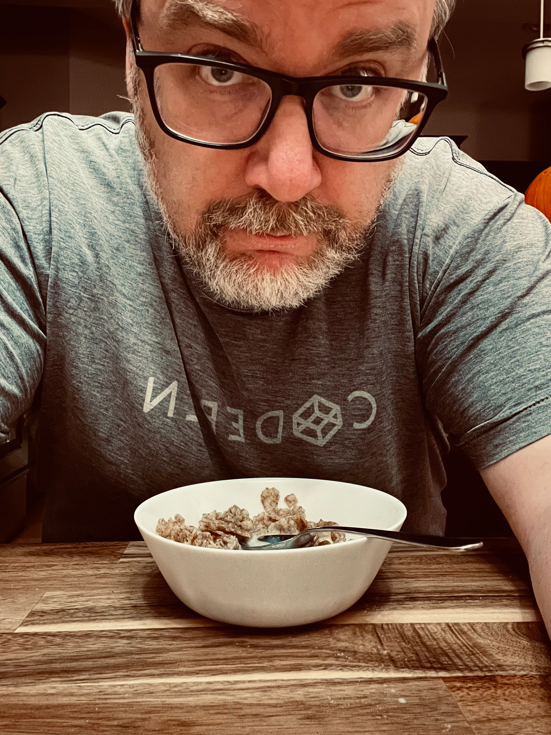 A man with glasses and a beard is sitting at a table with a bowl of cereal in front of him.