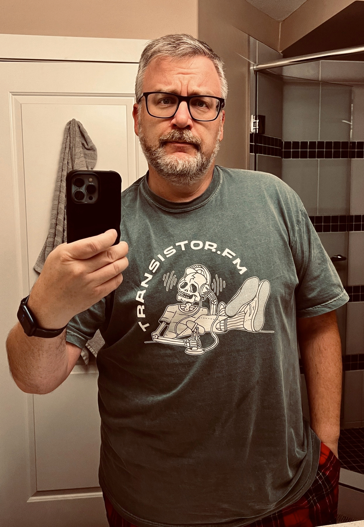 A man wearing glasses is taking a mirror selfie in a bathroom, wearing a T-shirt with a Transistor.fm design.